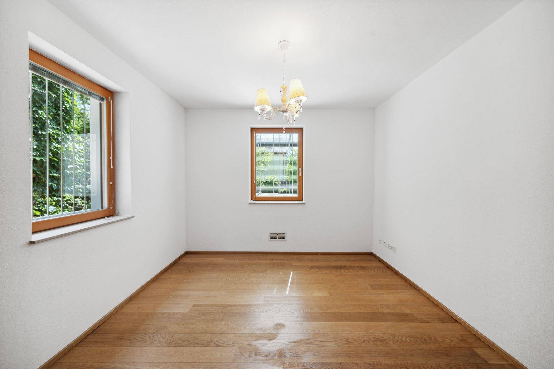 Leerer Raum mit Holzboden, zwei Fenstern und einem Kronleuchter an der Decke.