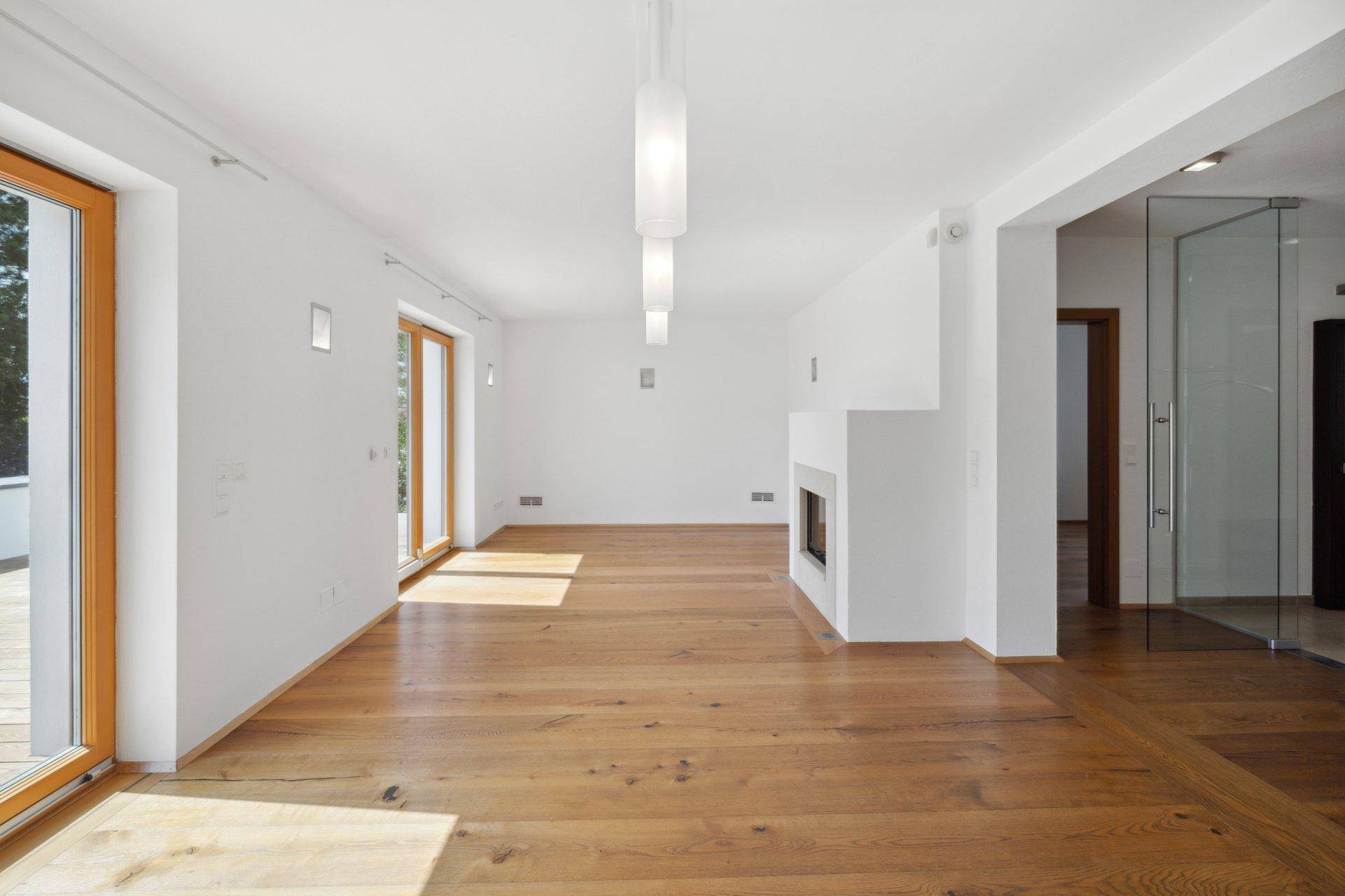 Helles, leeres Wohnzimmer mit Holzboden, modernem Kamin und großen Fenstern.