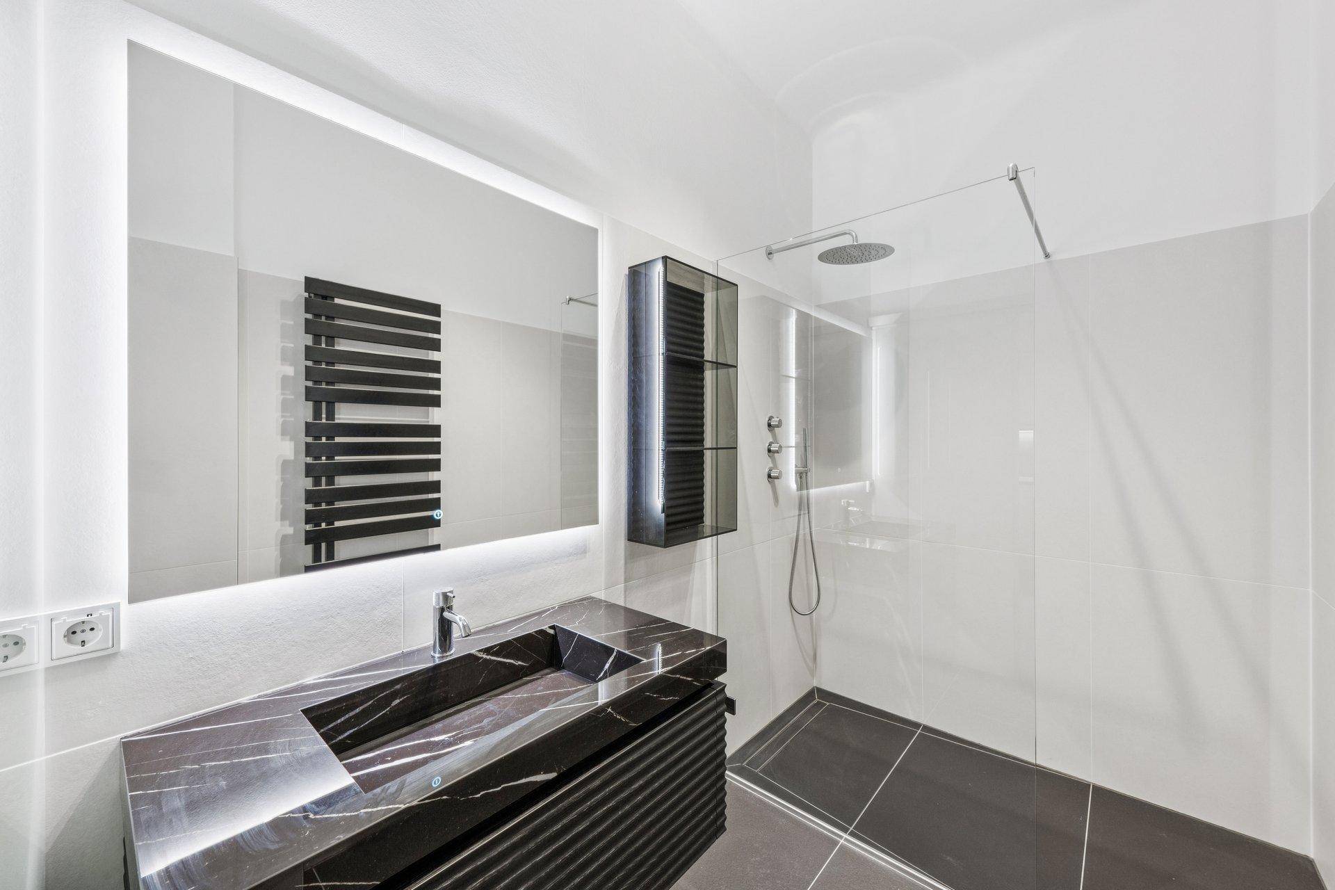 Modernes Badezimmer mit großer begehbarer Dusche, beleuchtetem Spiegel und dunklem Waschtisch.
