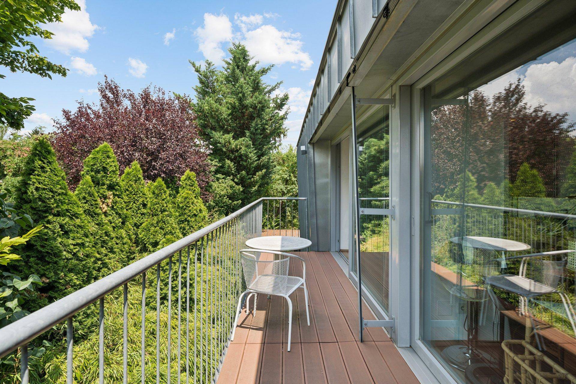 Kleiner Balkon mit Holzboden und Sitzgelegenheiten, umgeben von dichtem Grün und Bäumen.