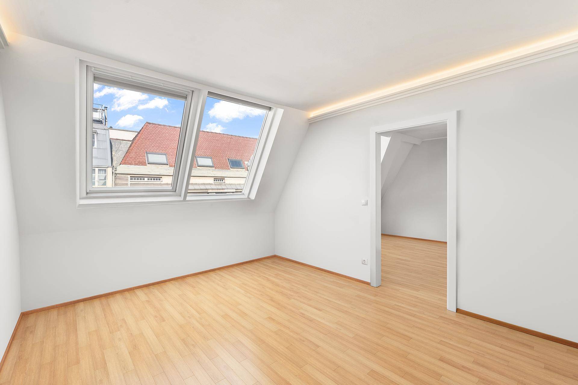 Helles Zimmer im Dachgeschoss mit Holzfußboden, Dachfenstern und indirekter Deckenbeleuchtung.