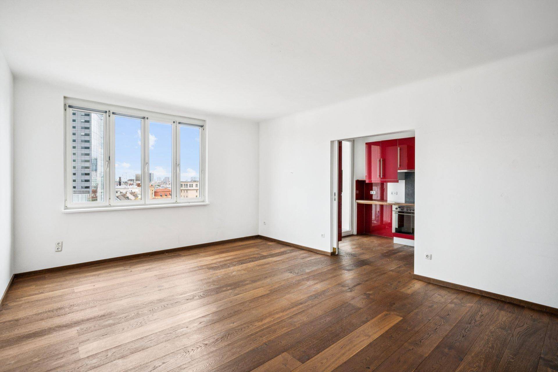Heller, leerer Raum mit Holzboden und großem Fenster, angrenzend an eine rote Küche.