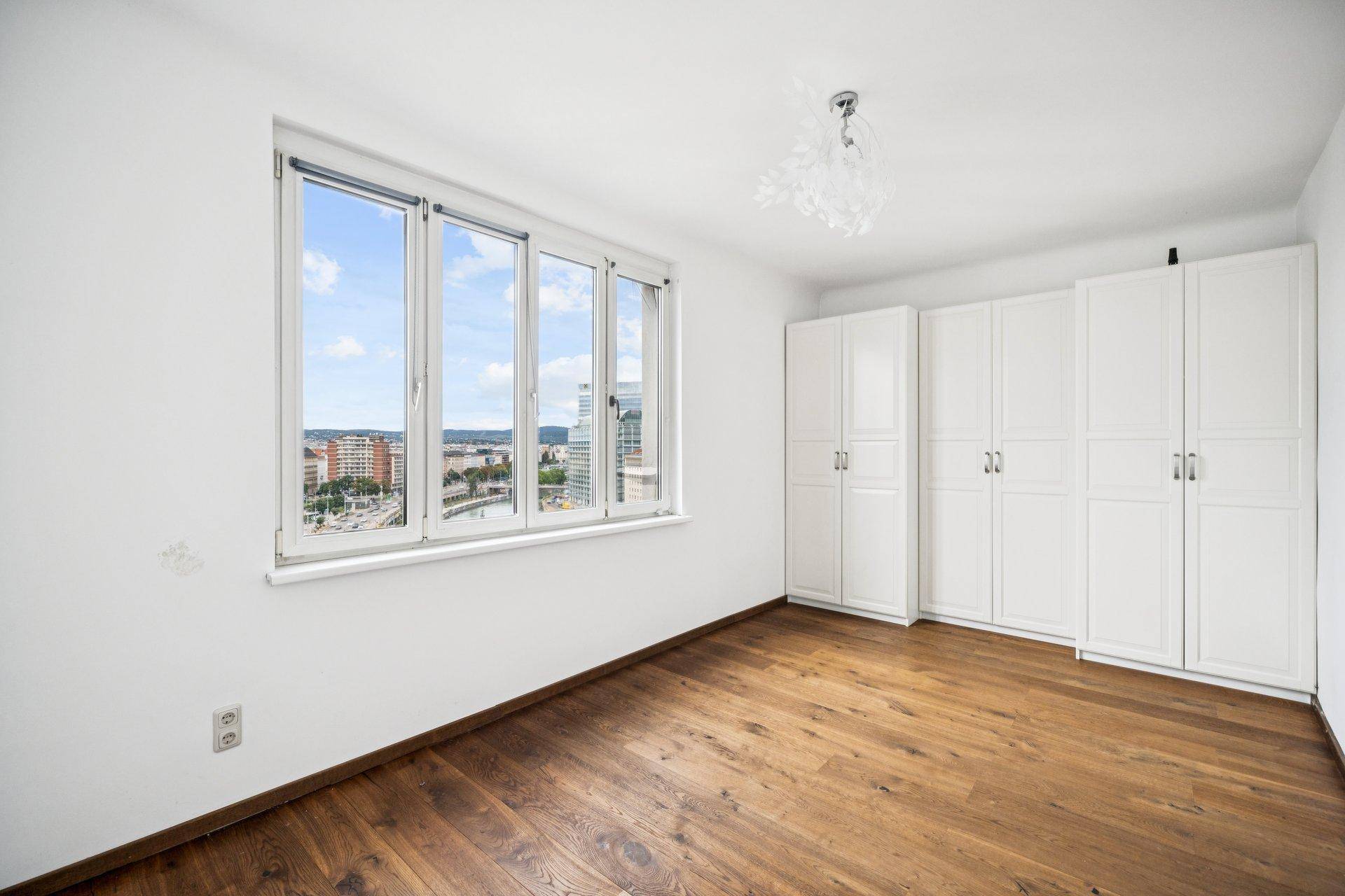 Geräumiges Schlafzimmer mit Holzboden, weißen Einbauschränken und Stadtblick.