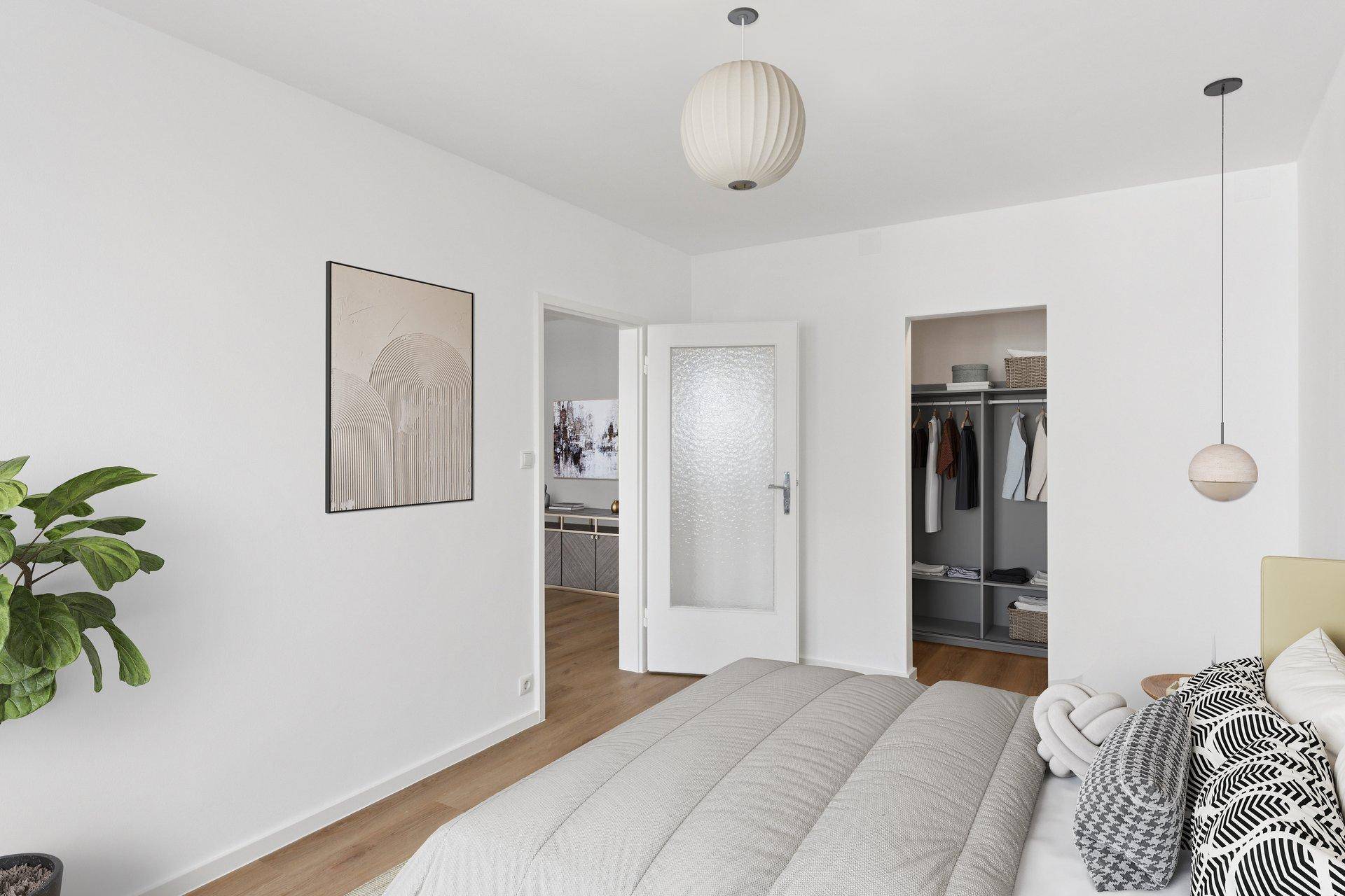 Schlafzimmer mit hellem Bett, Kleiderschrank und Zugang zu einem weiteren Raum.