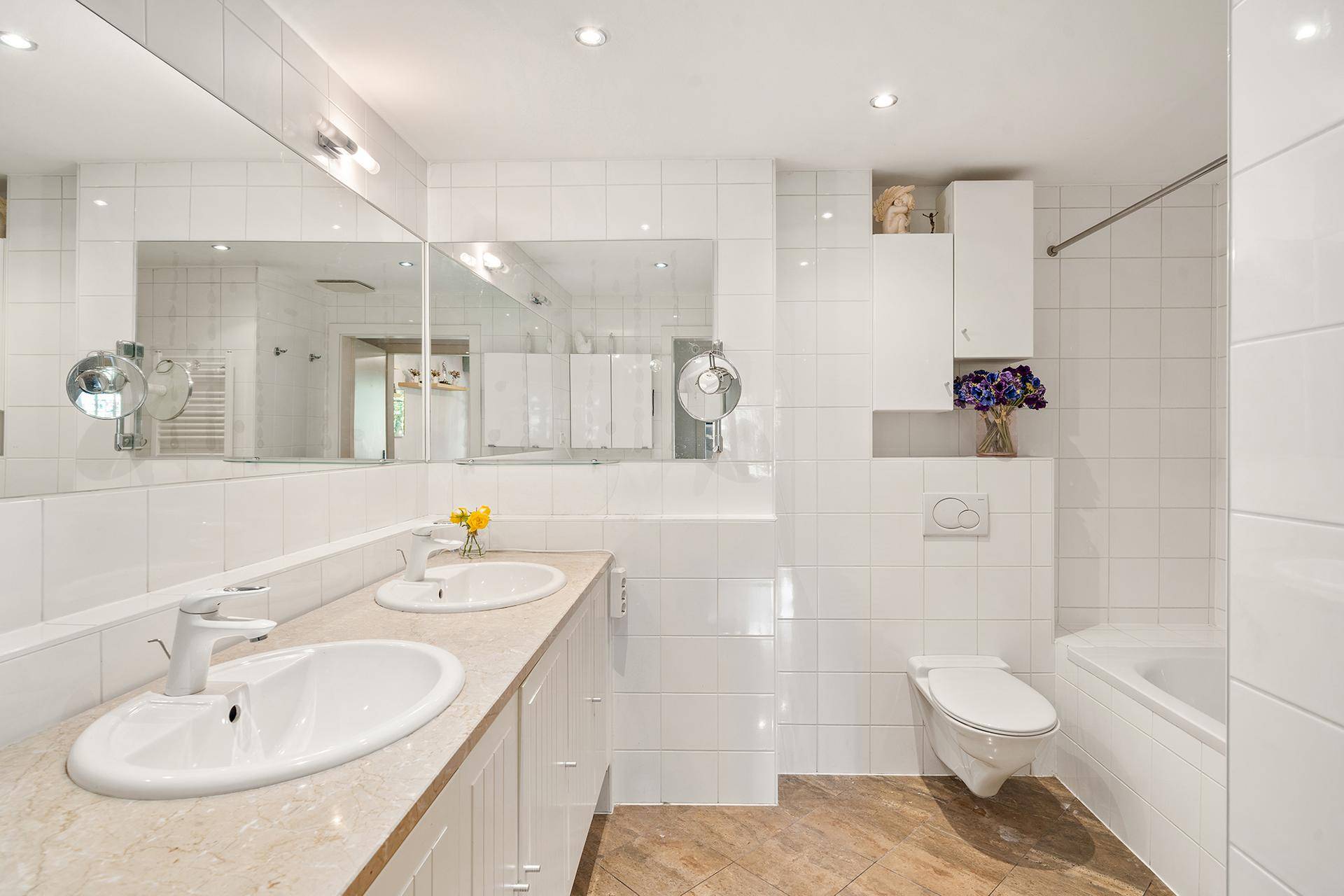Modernes Badezimmer mit weißen Fliesen, Doppelwaschbecken, großer Spiegelfläche und Badewanne.