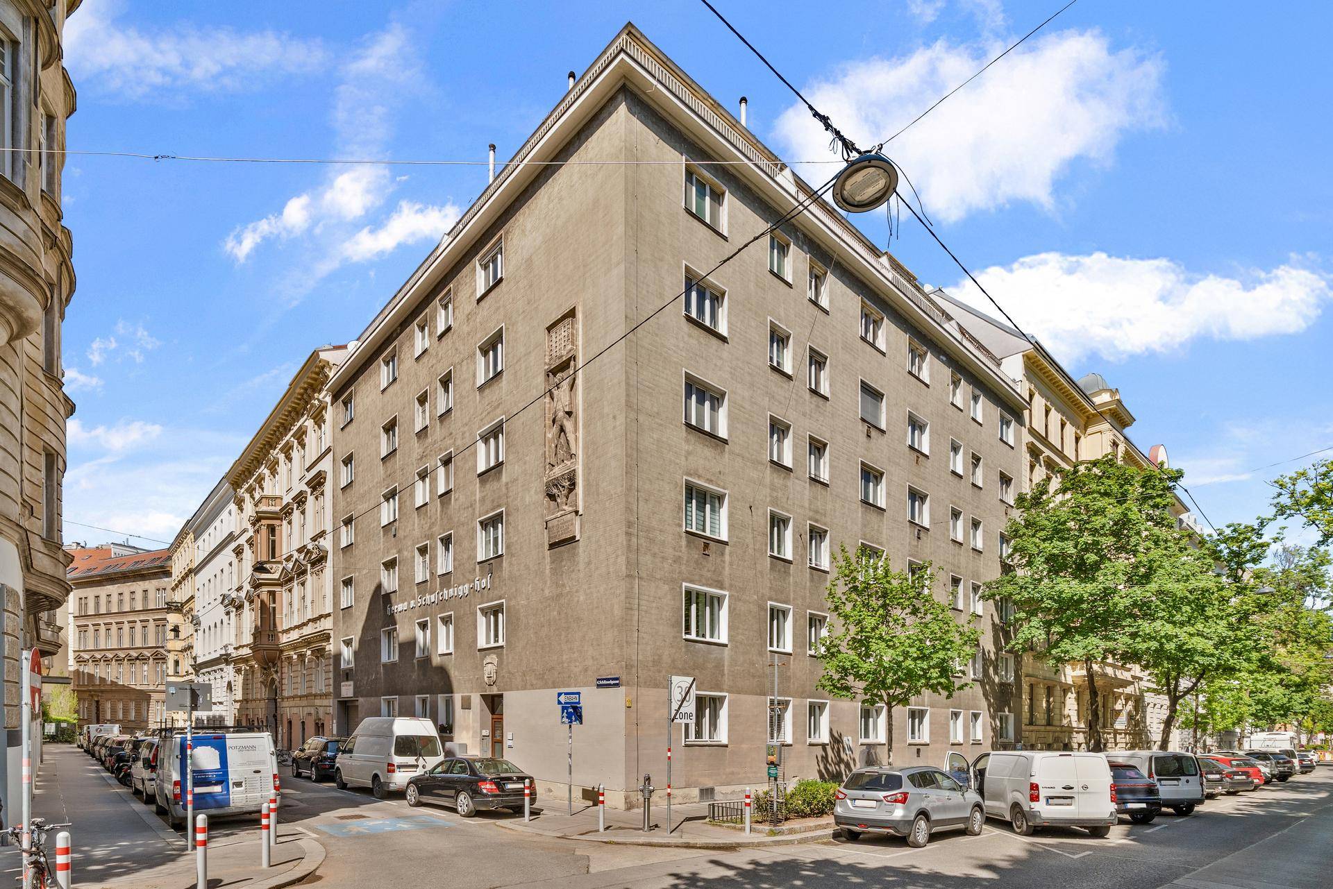 Eckgebäude in städtischer Umgebung mit mehreren Stockwerken und vielen Fenstern unter blauem Himmel.