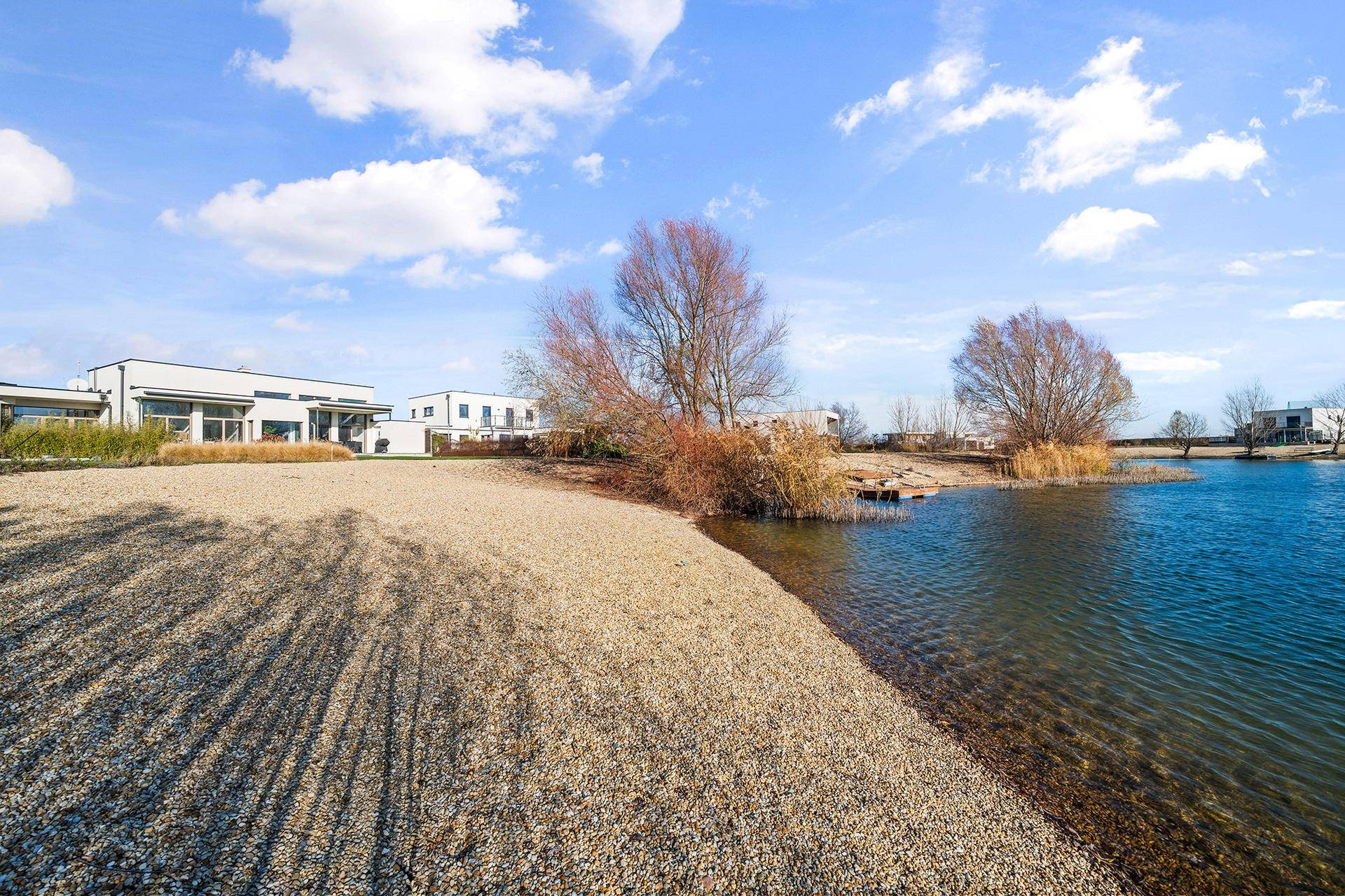 Kiesstrand am Seeufer mit Blick auf moderne Häuser und die umliegende Natur.