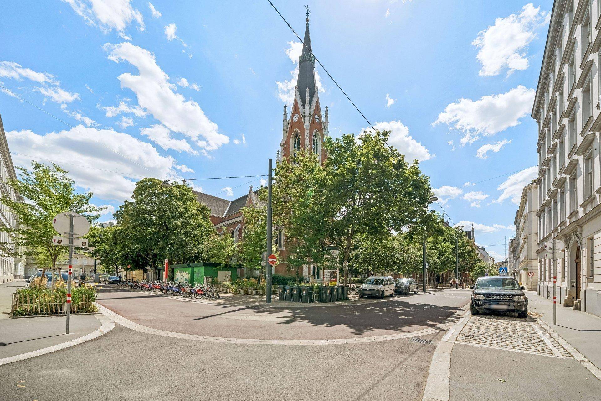 Straßenansicht mit historischer Kirche, Bäumen und geparkten Autos in urbaner Umgebung.