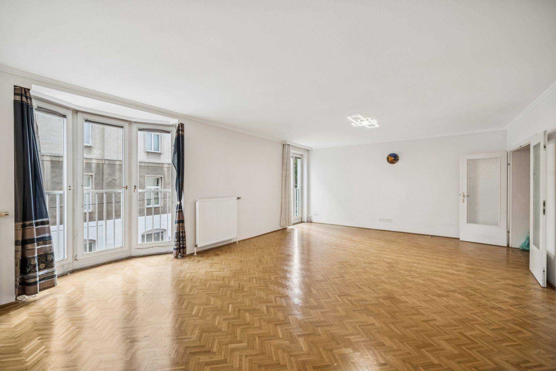 Heller Wohnbereich mit Fischgrätparkett, großen Fenstern und Zugang zu einem französischen Balkon.