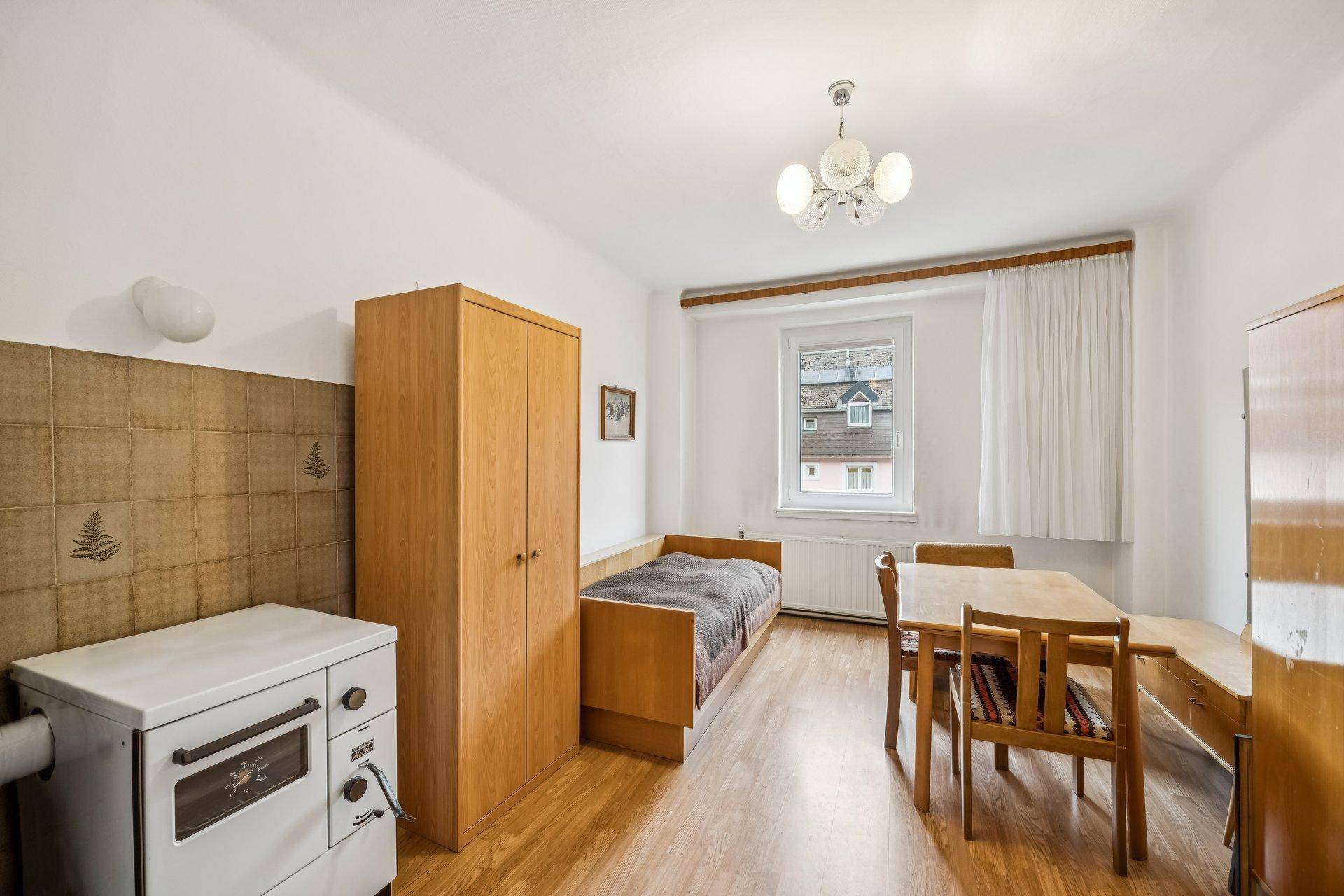 Einfaches Zimmer mit Einzelbett, Holzschrank, Esstisch und traditionellem Ofen.