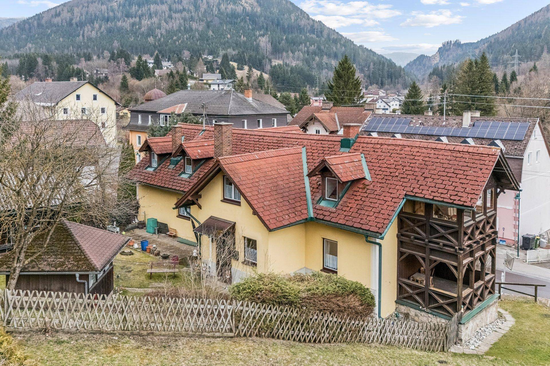 Traditionelles Haus mit rotem Ziegeldach und Holzbalkon, umgeben von grüner Natur.