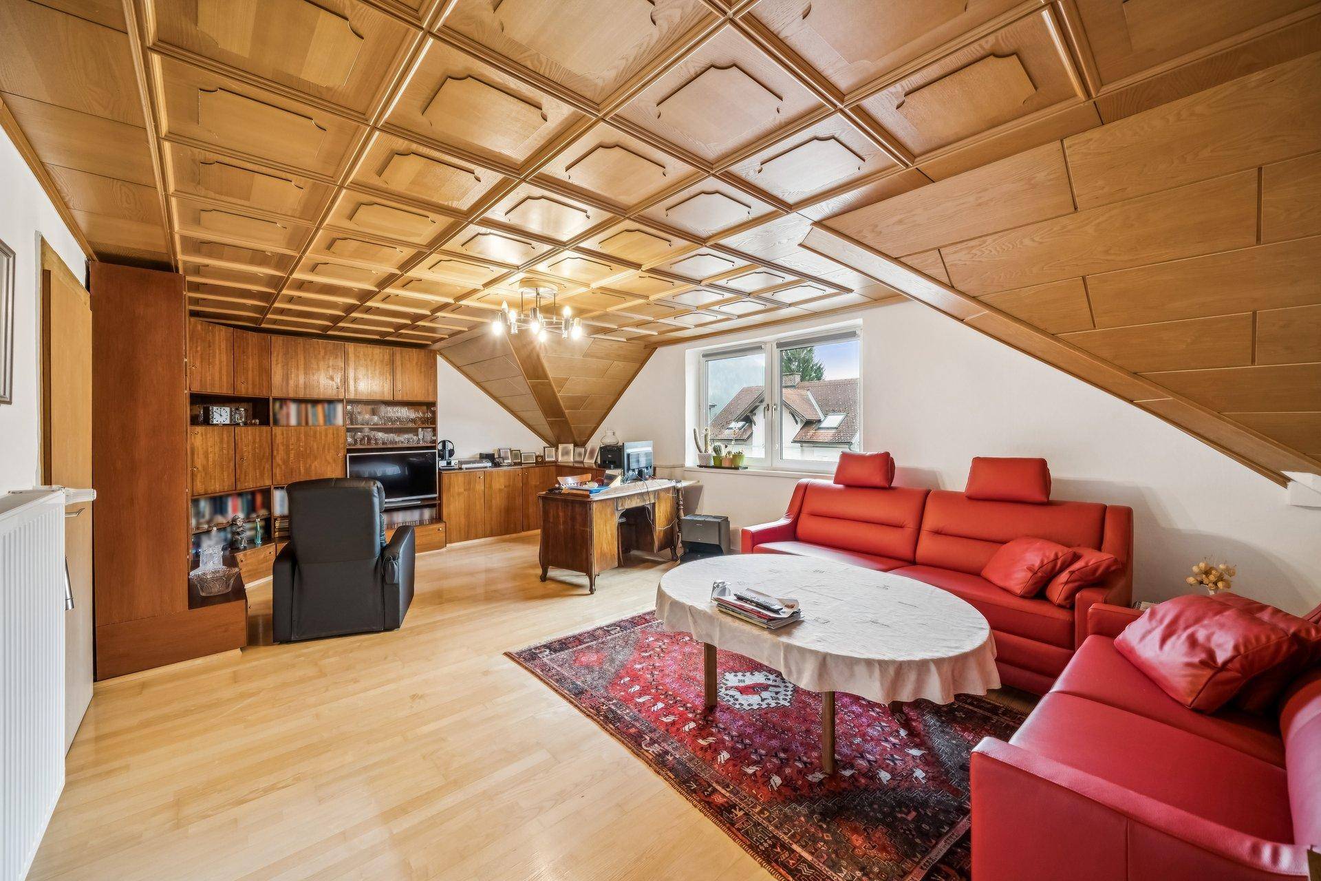 Gemütliches Wohnzimmer im Dachgeschoss mit Holzdecke, roten Sofas und einem Schreibtischbereich.