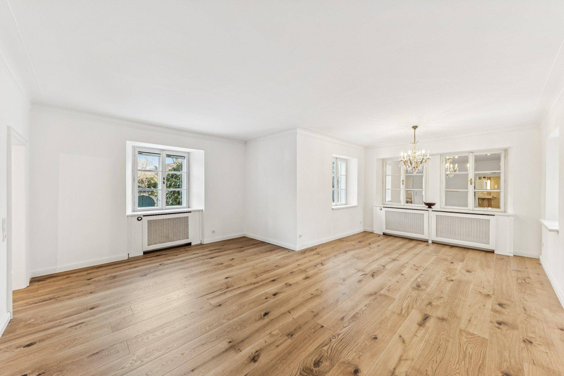 Geräumiger Raum mit Holzdielenboden, großen Fenstern und einem eleganten Kronleuchter.