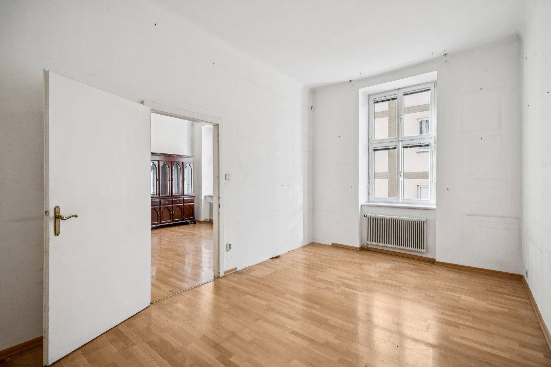 Helles Zimmer mit Parkettboden und einem Fenster, das zu einem weiteren Raum führt.