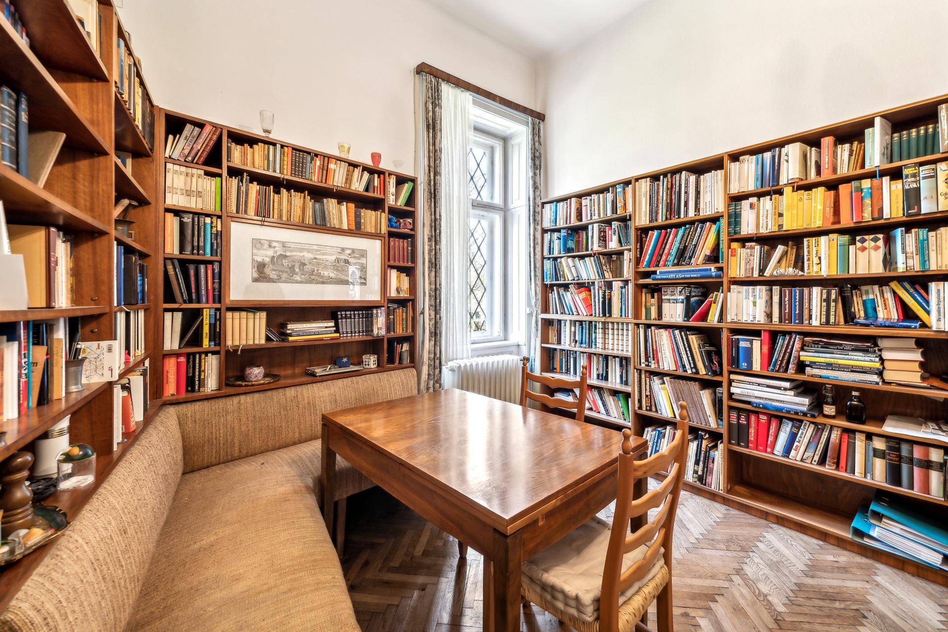 Helles Arbeitszimmer mit raumhohen Bücherregalen, Holztisch und Parkettboden.