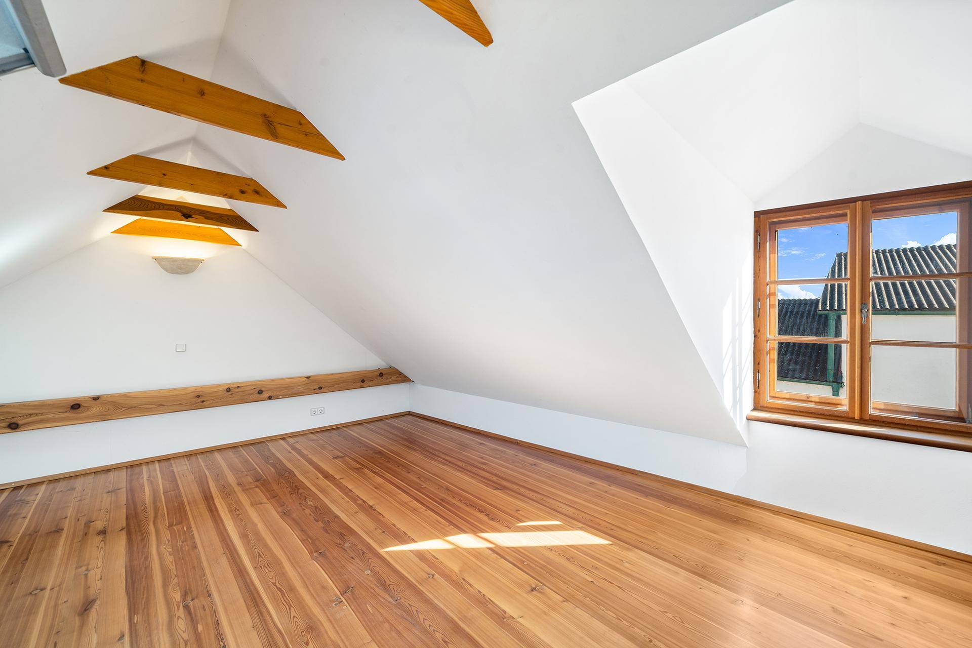 Helles Dachzimmer mit Holzboden, schrägen Wänden und einem Fenster mit Blick nach draußen.