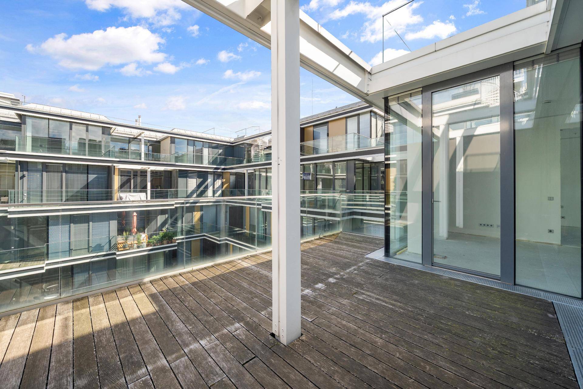 Große Holzterrasse mit Glasgeländer und Blick auf die moderne Wohnanlage.