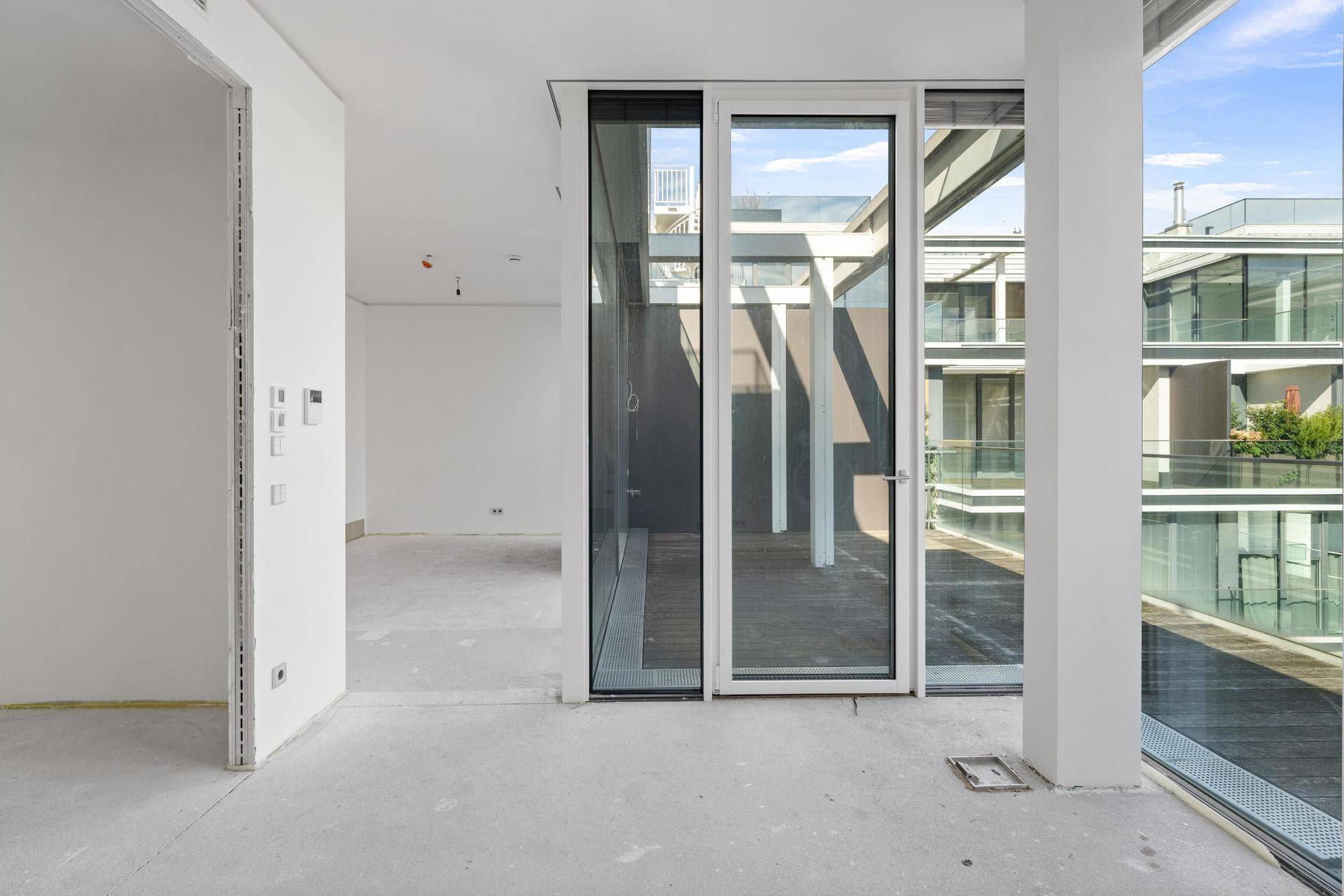 Geräumiger Innenraum im Rohbauzustand mit Betonboden und Zugang zur Terrasse.