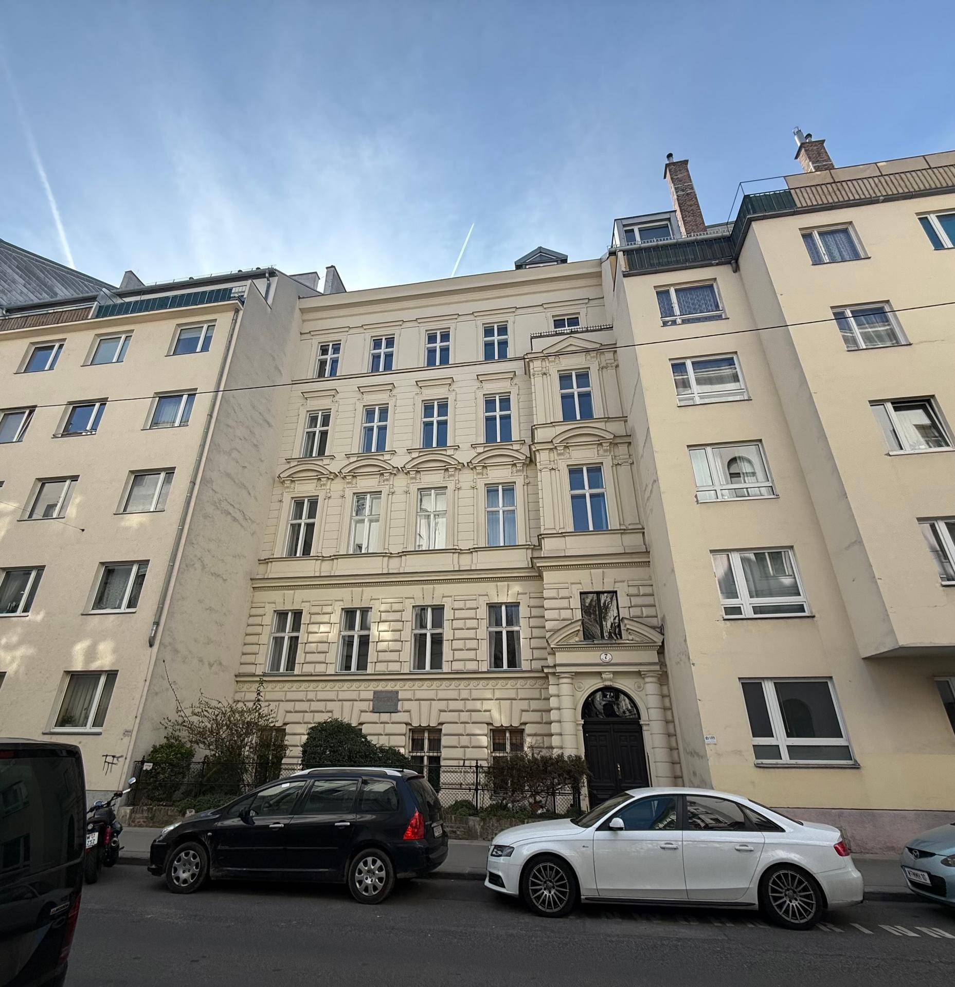 Historische Gebäudefassade an einer Straße mit geparkten Autos unter blauem Himmel.