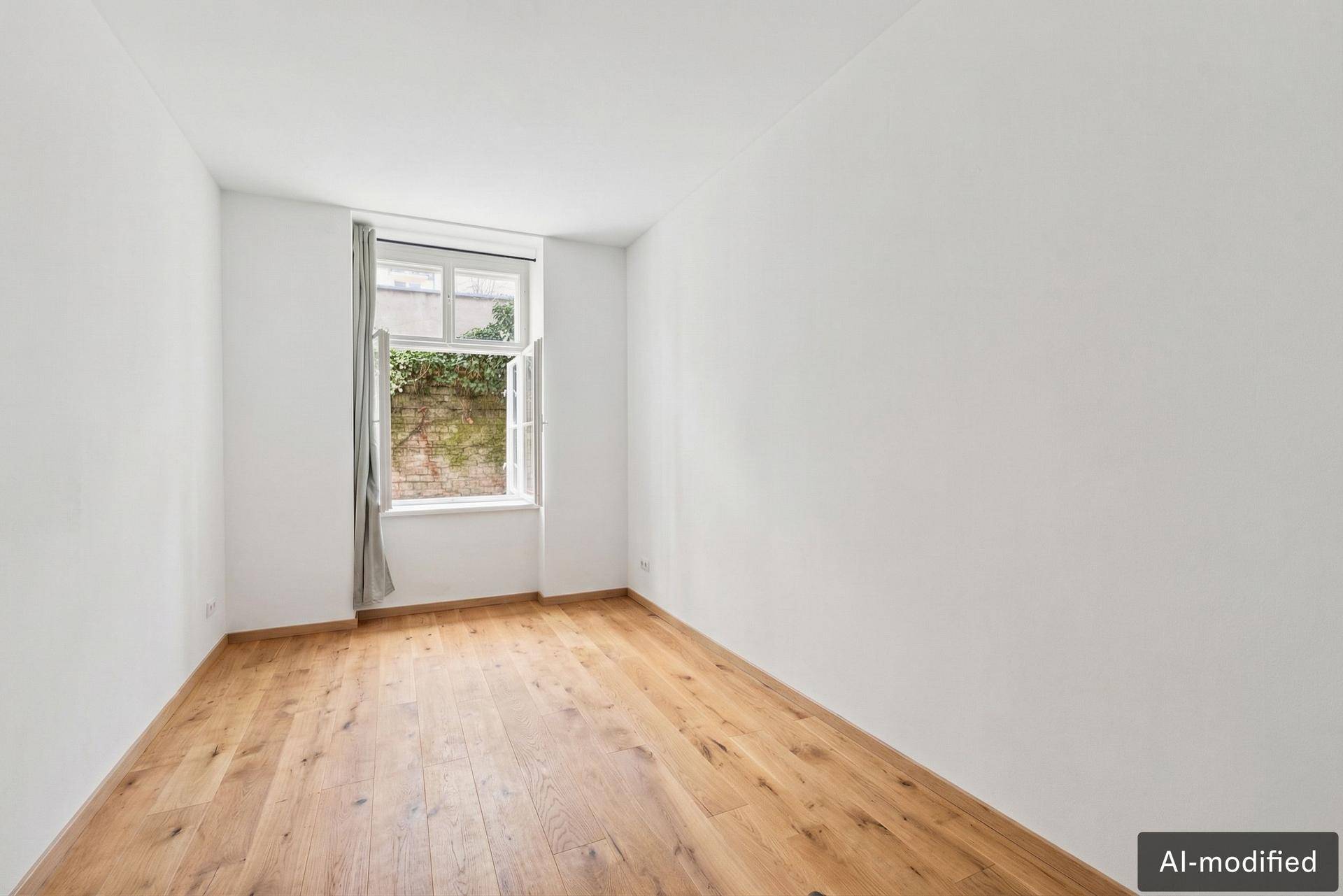 Leeres Zimmer mit Holzboden und Fenster mit Blick ins Grüne.