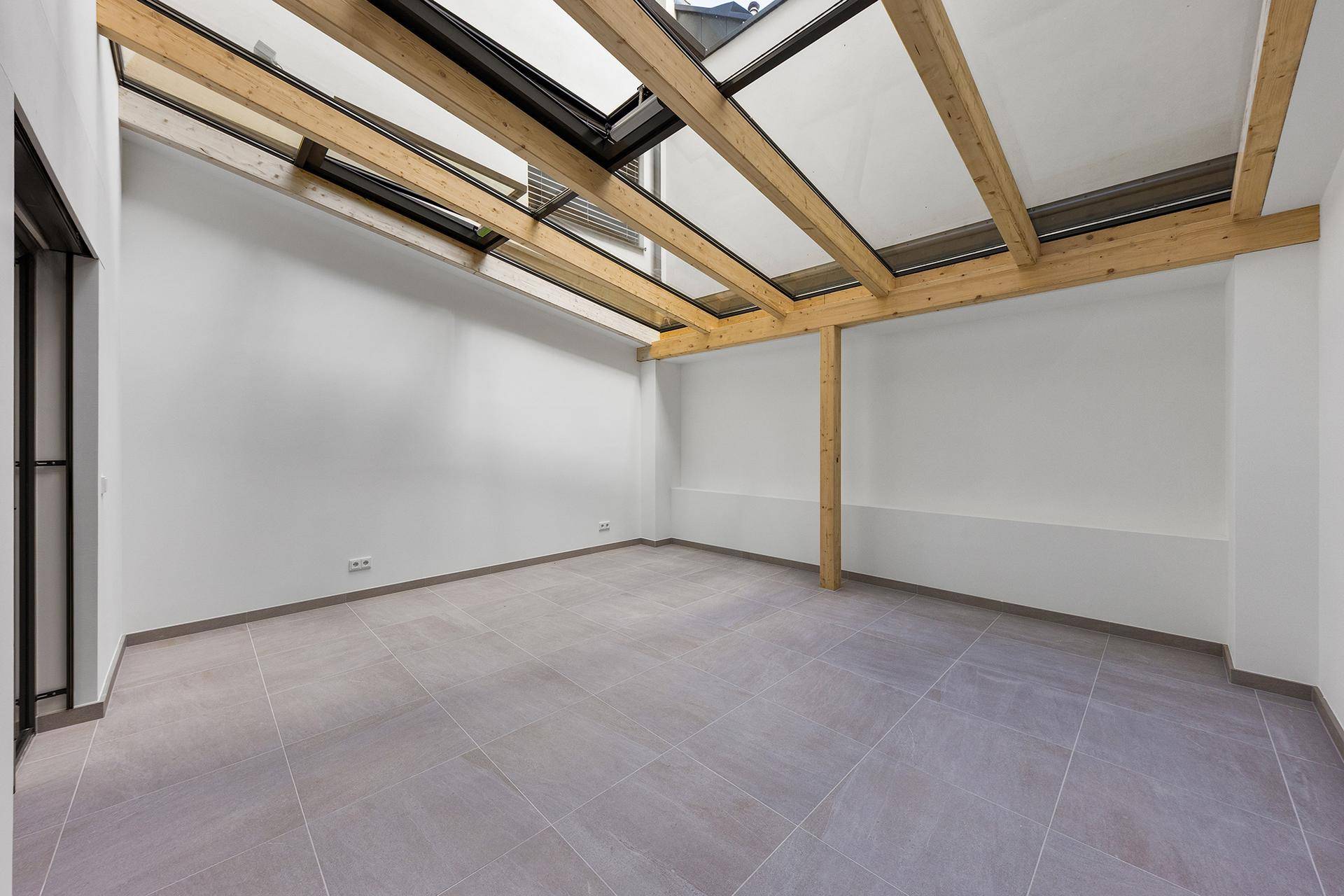 Heller Raum mit Fliesenboden und großer Dachverglasung mit sichtbaren Holzbalken.