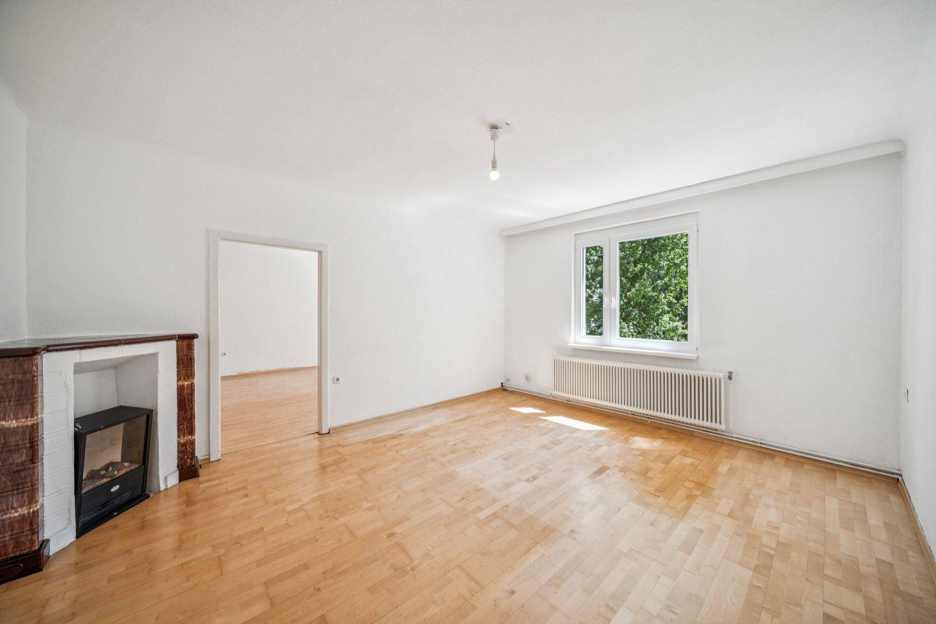 Geräumiges, unmöbliertes Wohnzimmer mit Parkettboden und einer Kaminattrappe.