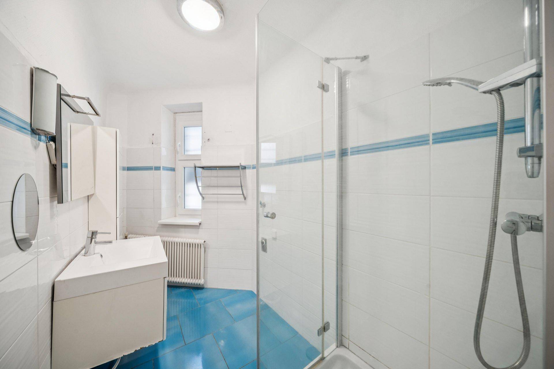 Badezimmer mit blauen Bodenfliesen, Duschkabine und Fenster für Tageslicht.