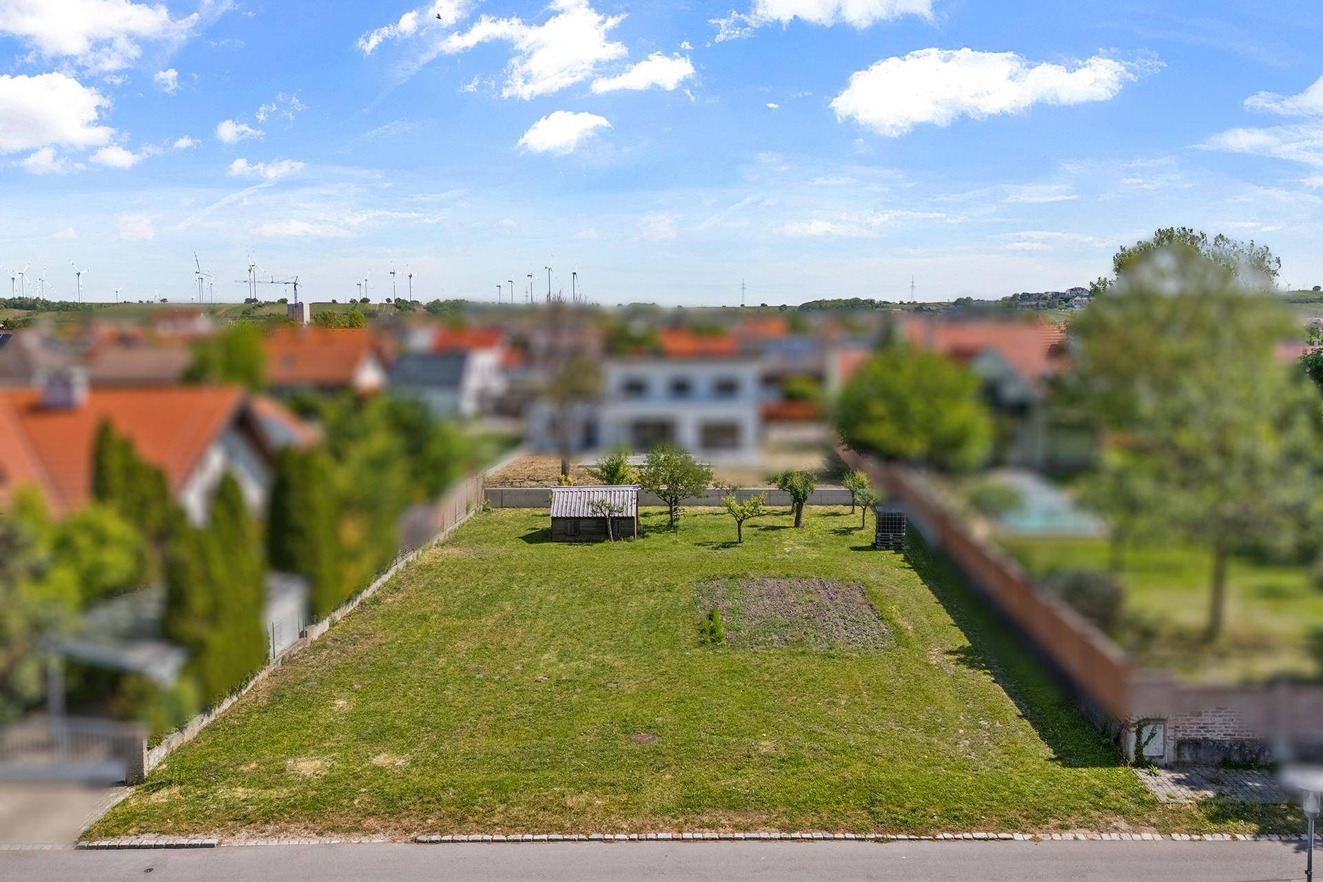 Luftaufnahme eines Grundstücks mit Bäumen und Schuppen, umgeben von Häusern und Windrädern in der Ferne.