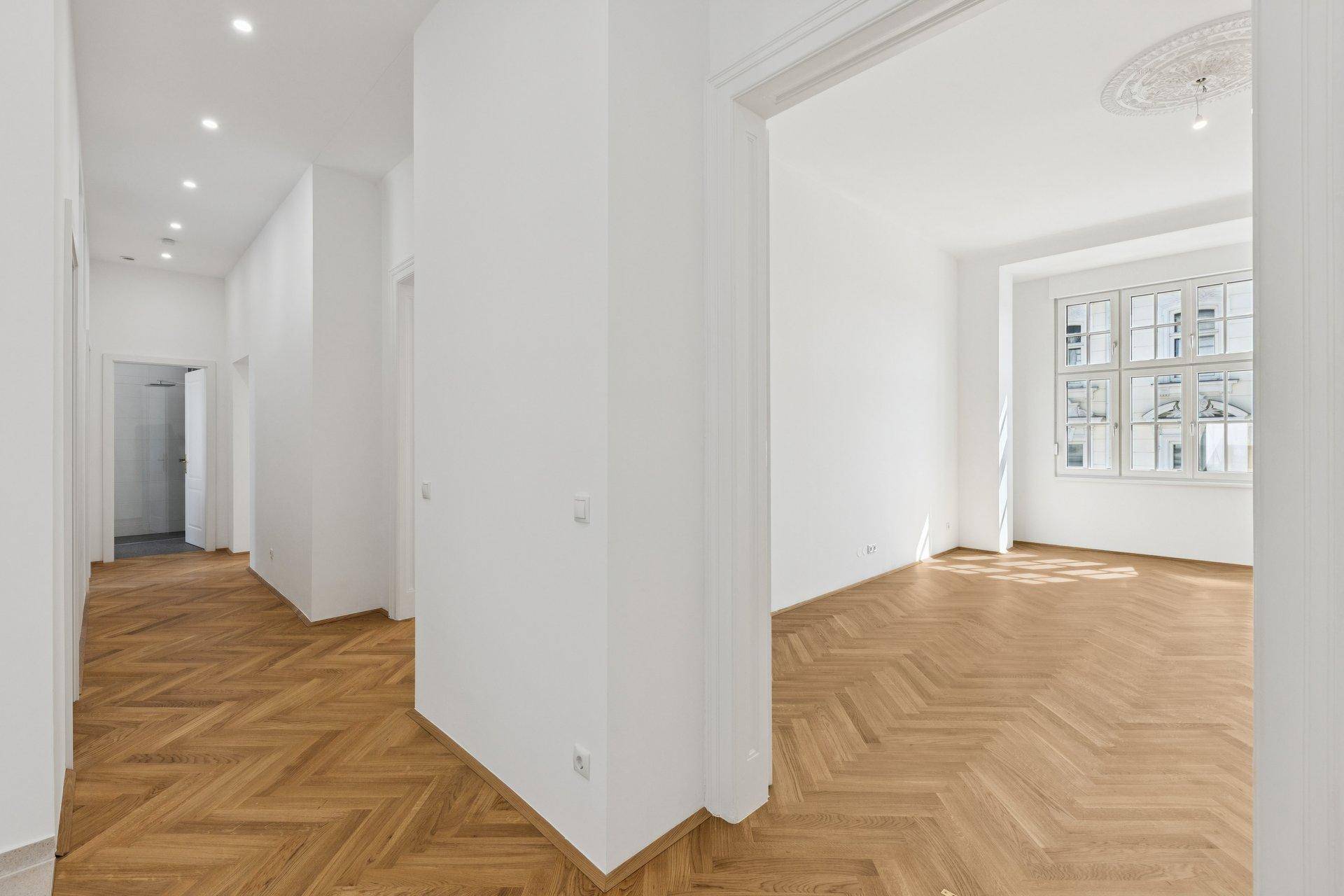 Heller Flur mit Fischgrätparkett und Blick in ein weiteres Zimmer mit Stuckdecke.