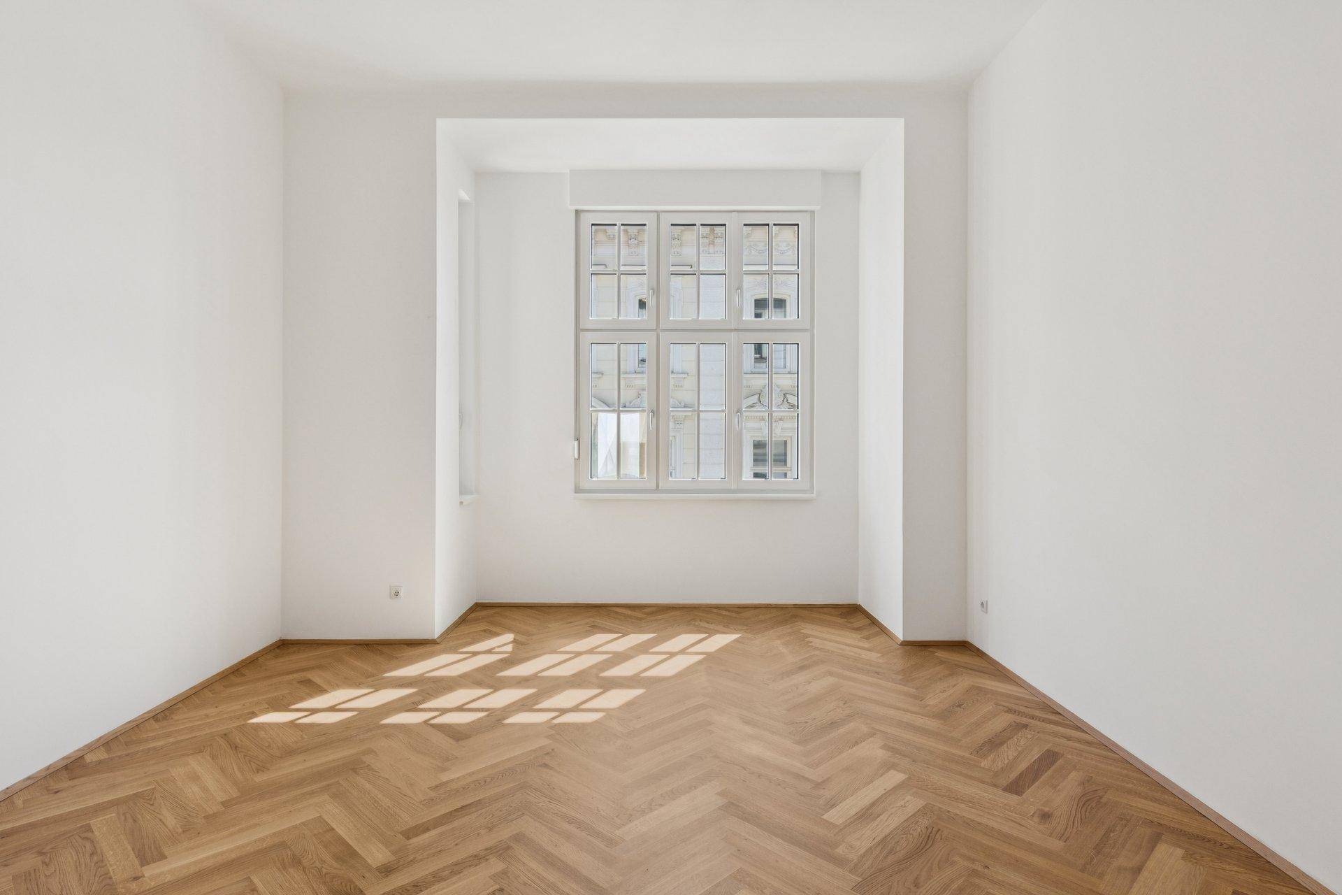 Helles Zimmer mit Fischgrätparkett und einem großen Fenster, das den Raum mit Sonnenlicht füllt.