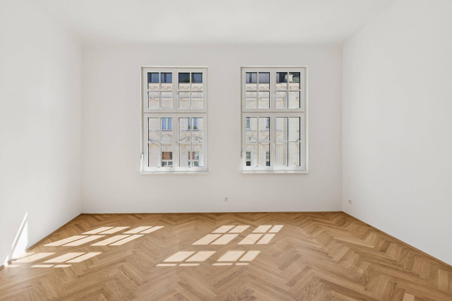 Geräumiges Zimmer mit Fischgrätparkett und zwei großen Fenstern für viel Tageslicht.