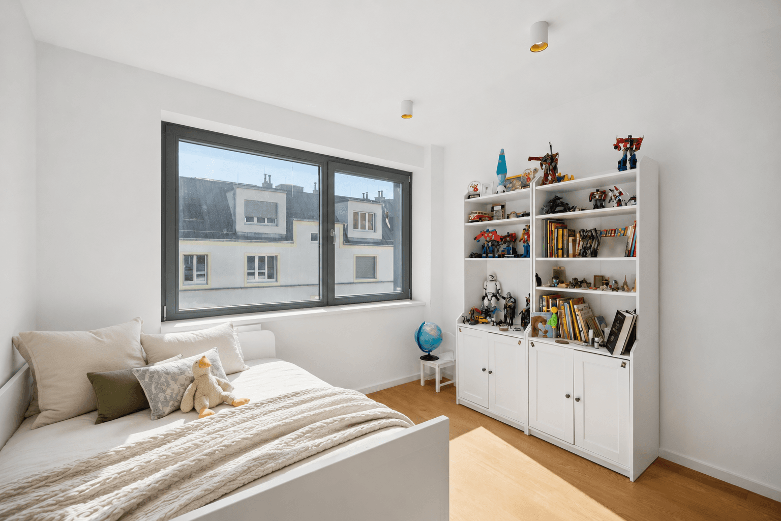 Kinderzimmer mit Bett, Bücherregal und großem Fenster mit Blick auf die Stadt.