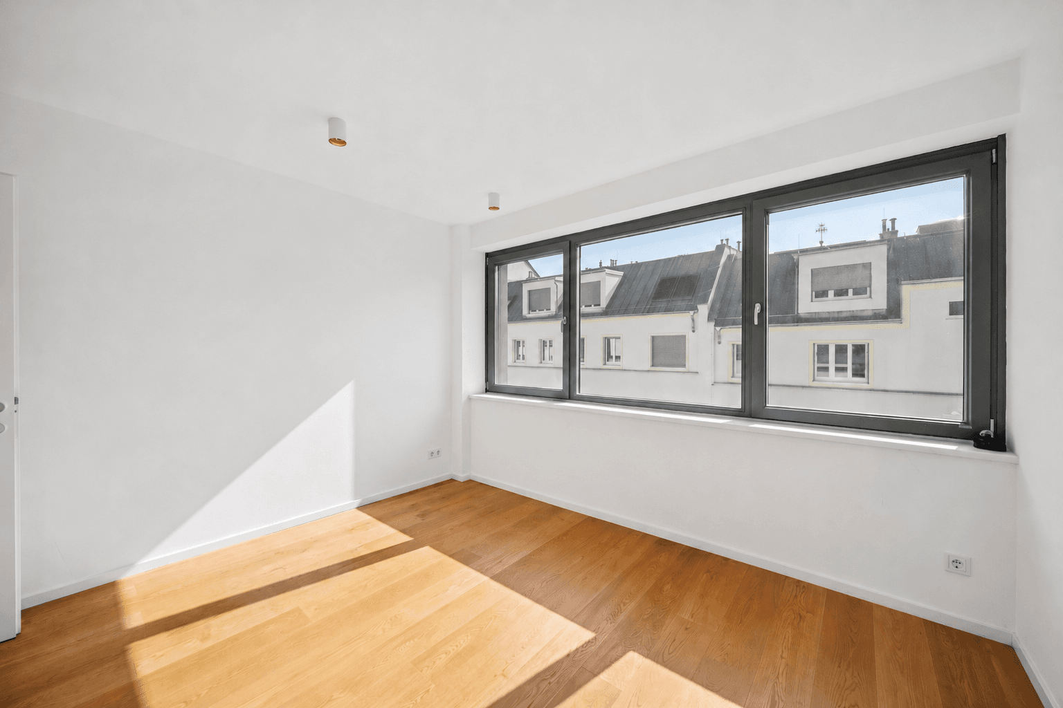 Leeres Zimmer mit Holzboden, großen Fenstern und Blick auf die umliegenden Dächer.