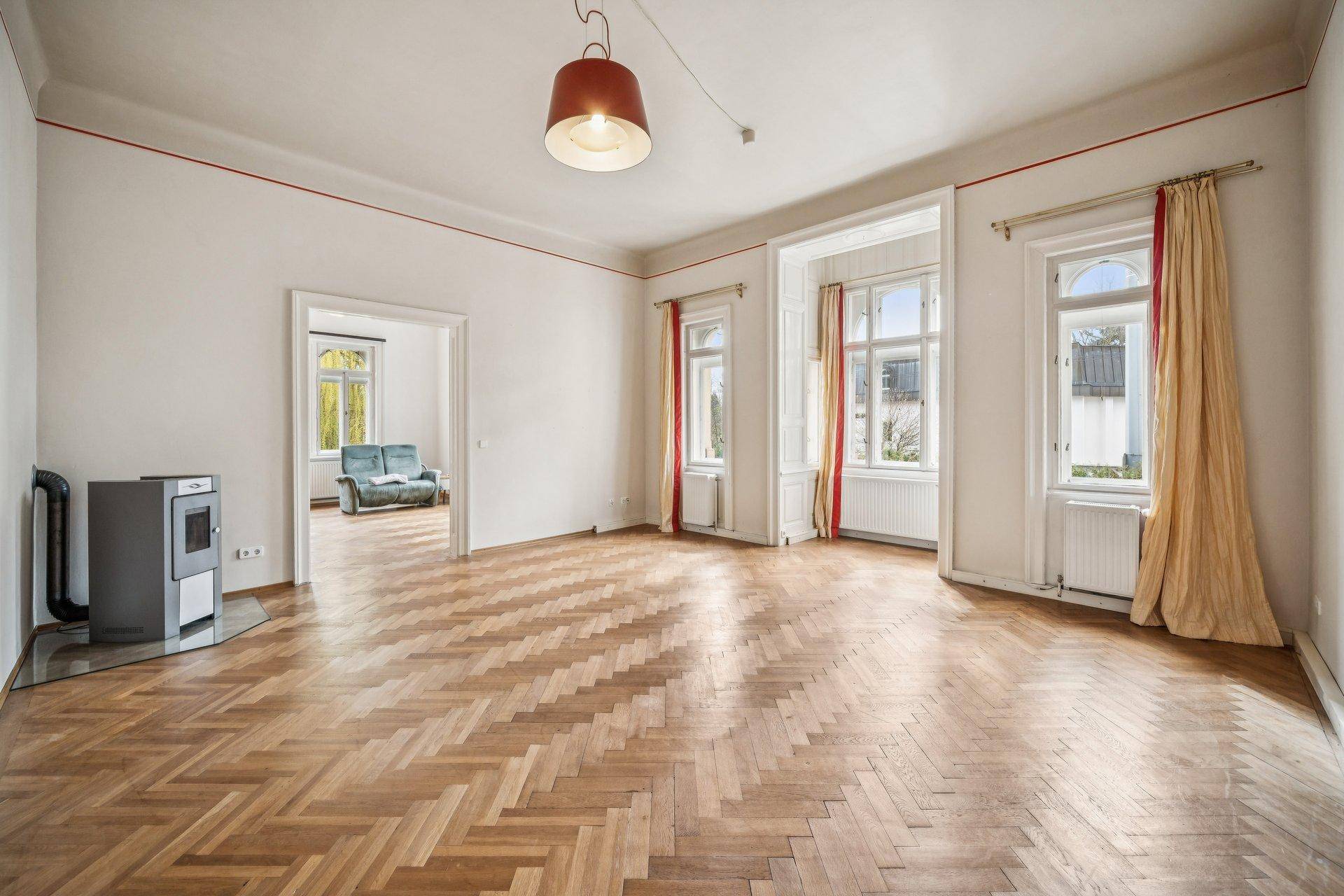 Geräumiges Wohnzimmer mit Fischgrätparkett, hohen Fenstern und einem modernen Holzofen.
