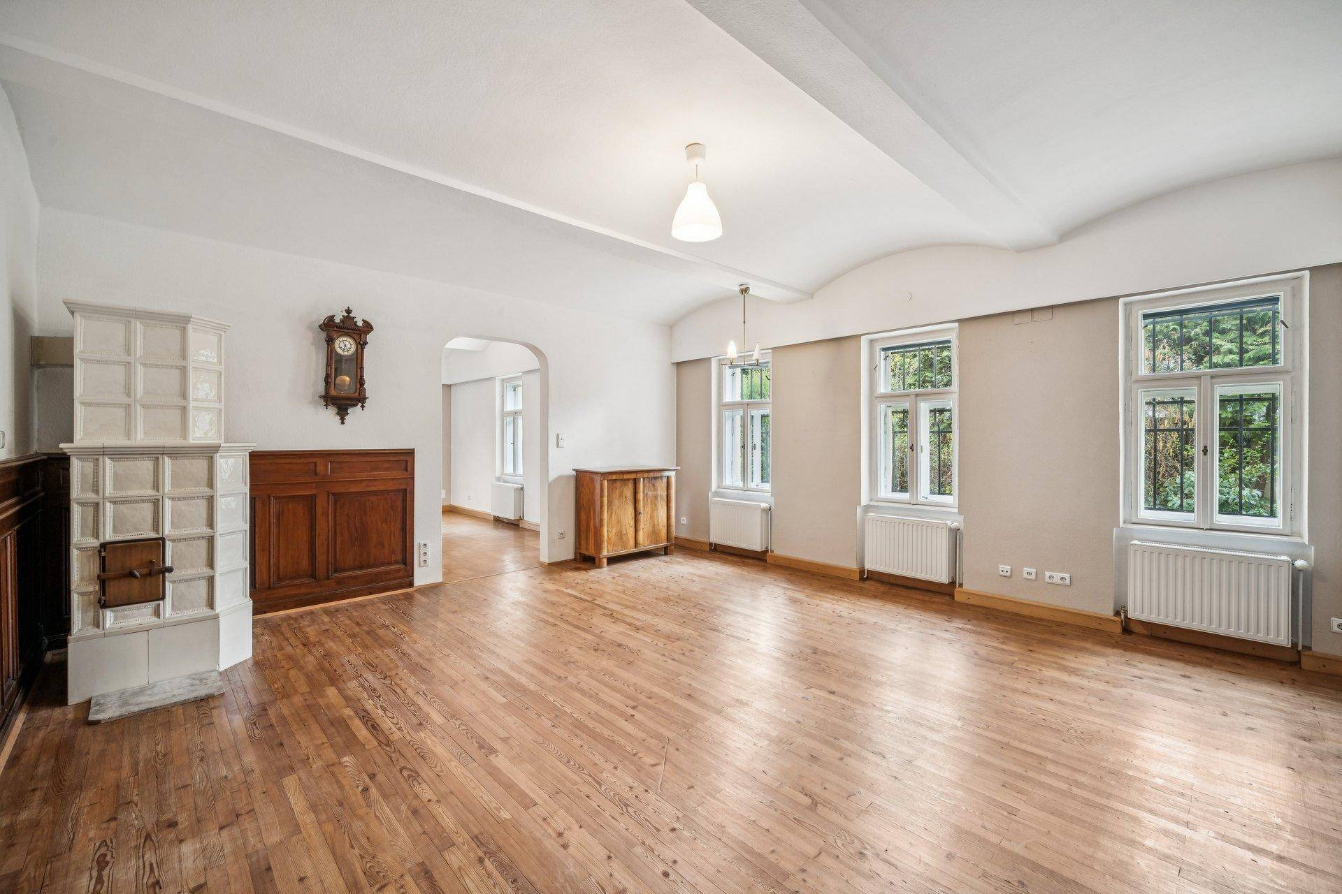 Heller Wohnraum mit Dielenboden, Kachelofen und Blick ins Grüne durch hohe Fenster.