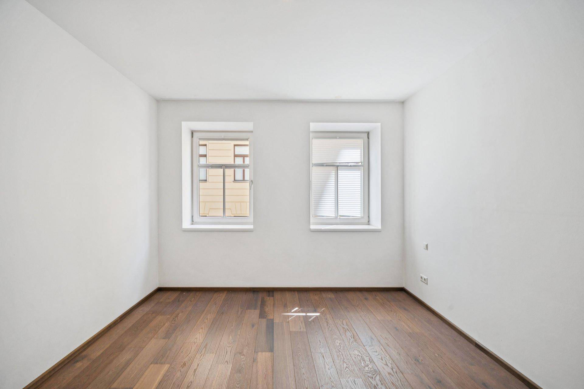Heller, leerer Raum mit Holzboden und zwei Fenstern, ideal als Schlaf- oder Arbeitszimmer.