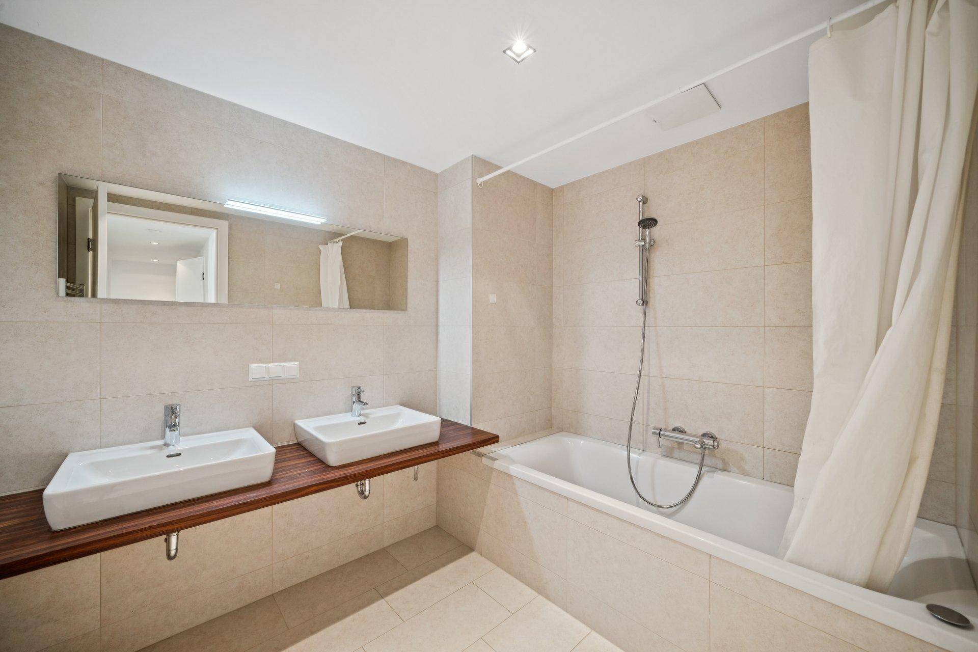 Helles Badezimmer mit beigefarbenen Fliesen, Doppelwaschbecken auf Holzplatte und Badewanne mit Duschvorhang.