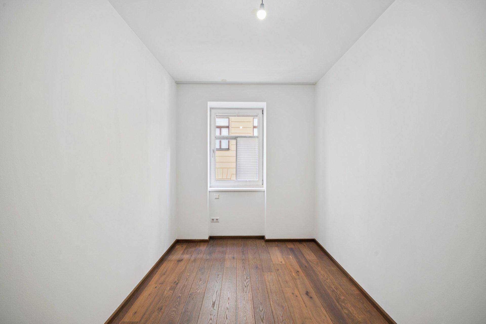 Kleiner, heller Raum mit Holzboden und einem Fenster, geeignet als Kinder- oder Gästezimmer.