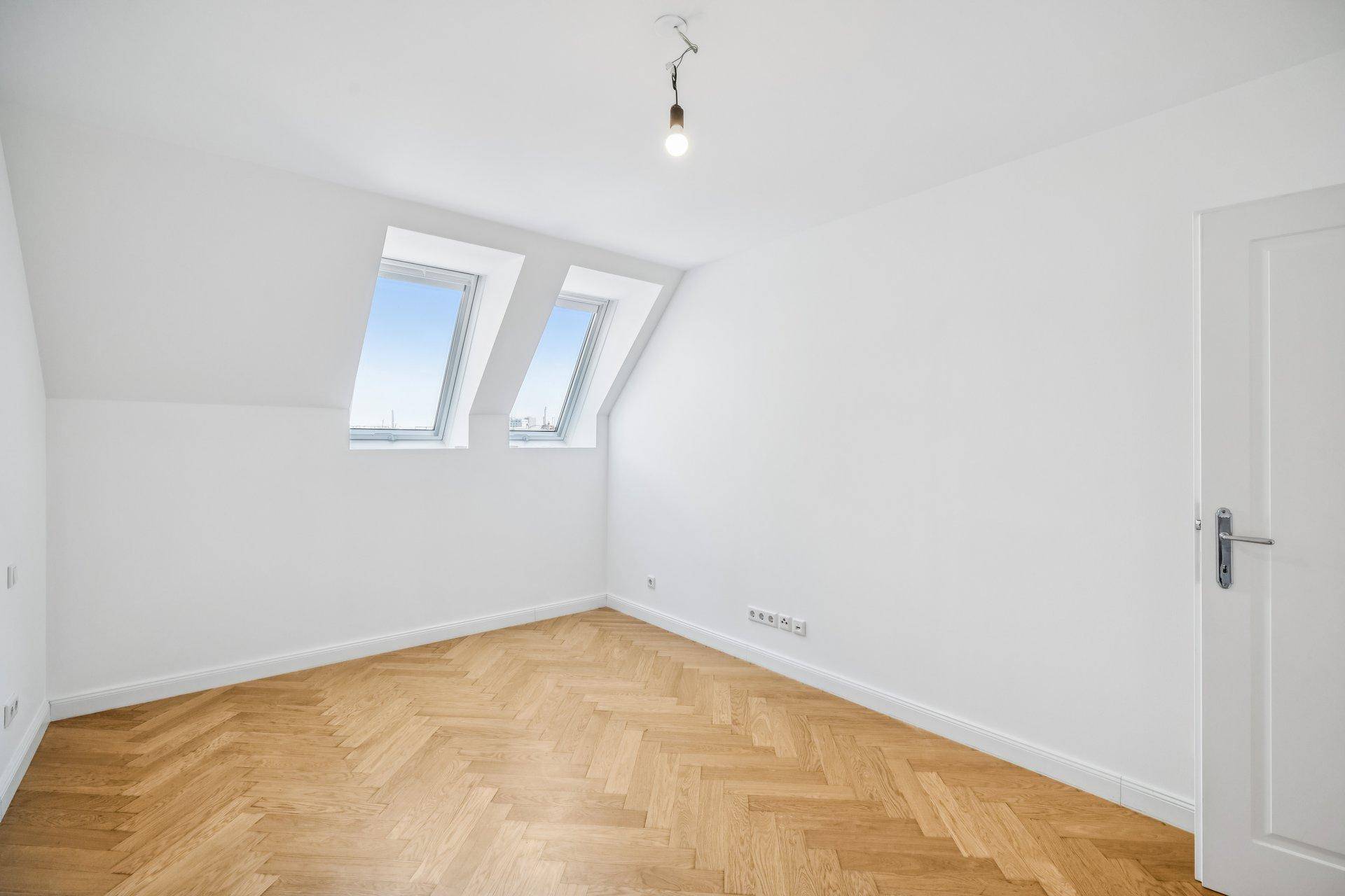 Leeres Schlafzimmer im Dachgeschoss mit schrägen Wänden, Dachfenstern und Fischgrätparkett.