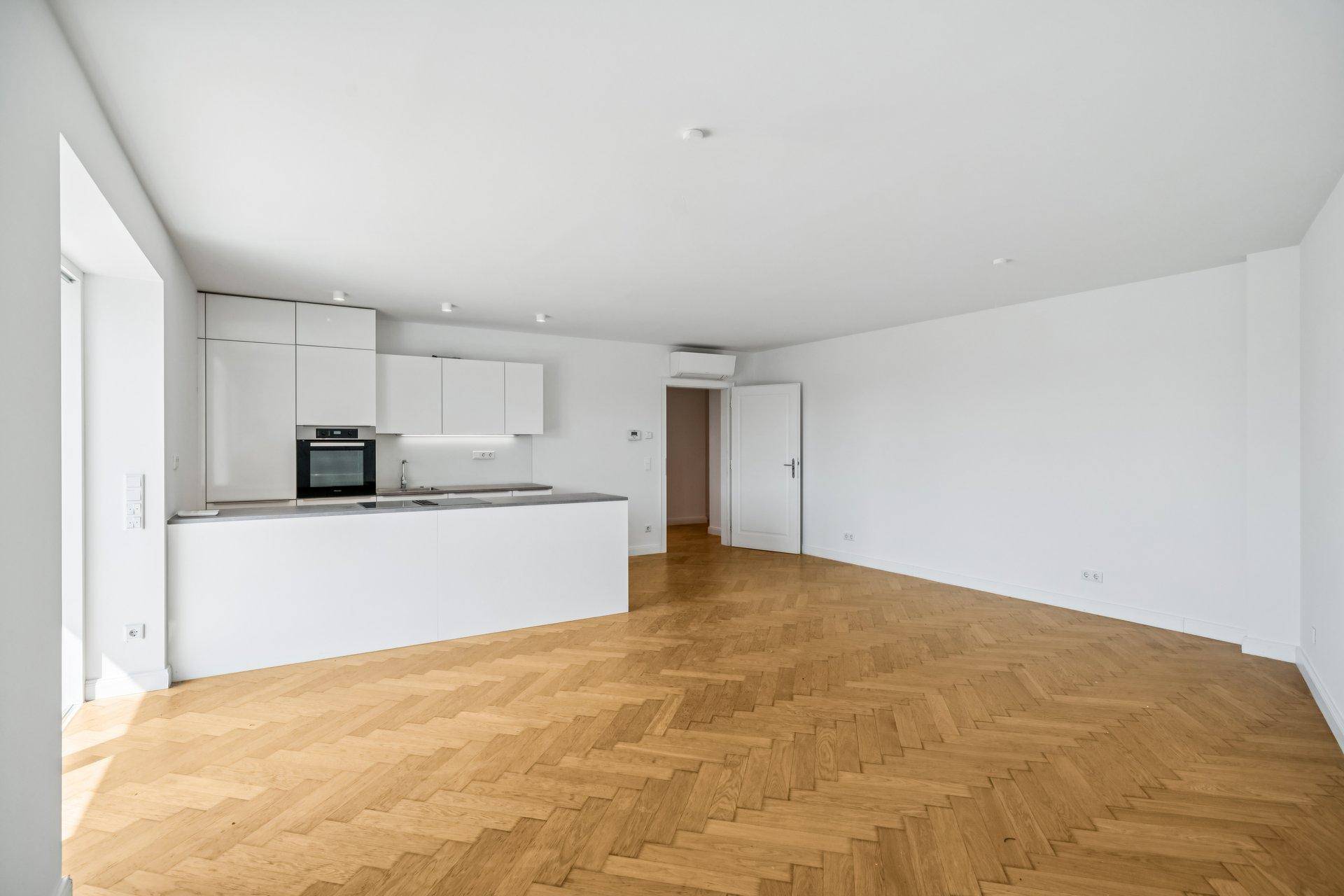 Geräumiger, leerer Wohnbereich mit offener Küche und elegantem Fischgrätparkettboden.