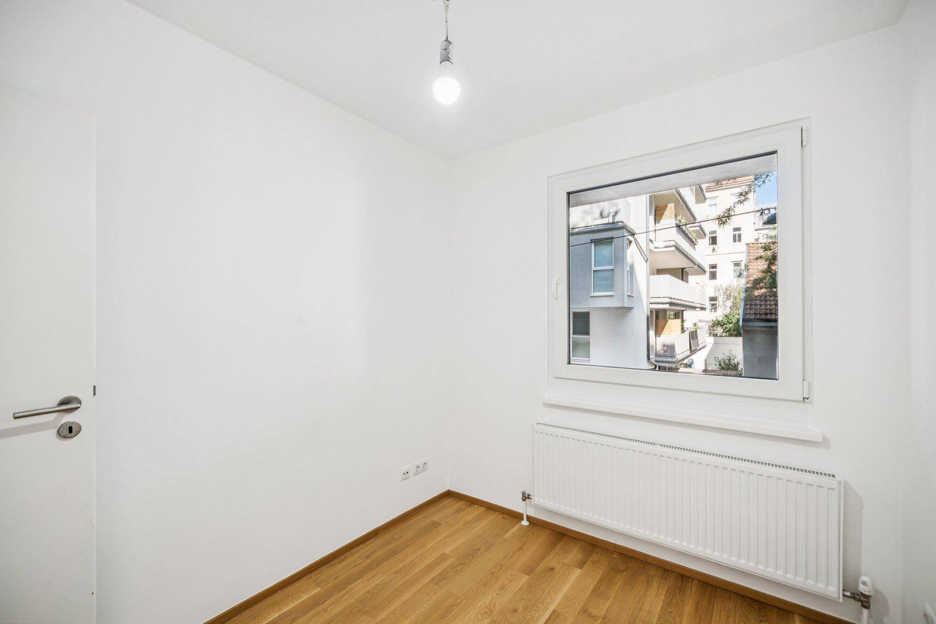 Kleinerer Raum mit Holzboden, weißen Wänden und einem Fenster mit Blick auf die Umgebung.