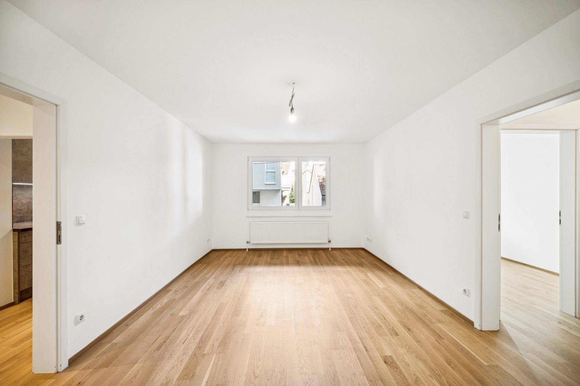 Leerer, heller Raum mit Parkettboden und einem Fenster, das viel Tageslicht hereinlässt.