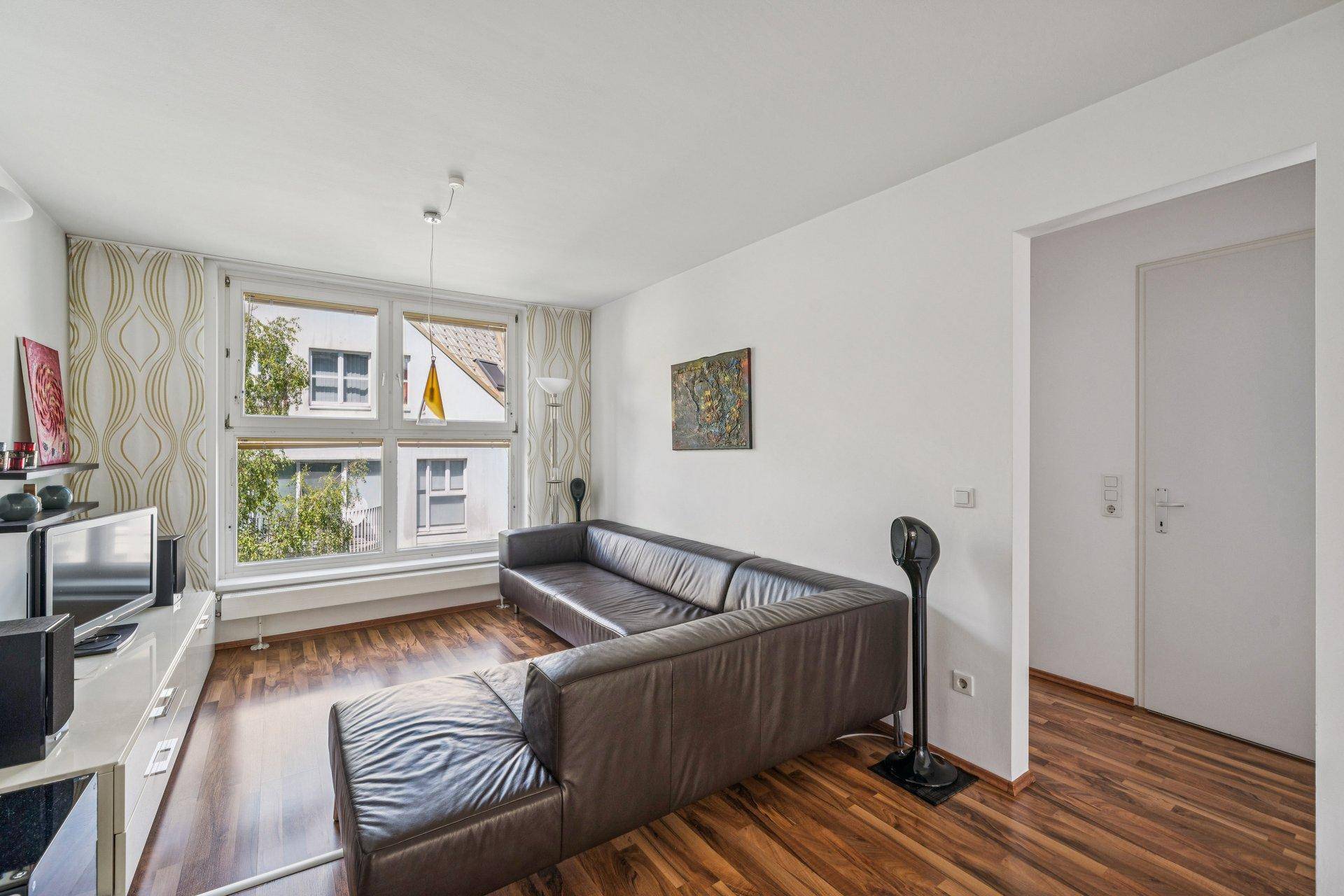 Gemütliches Wohnzimmer mit braunem Ecksofa, TV-Möbel und großem Fenster mit Blick ins Grüne.