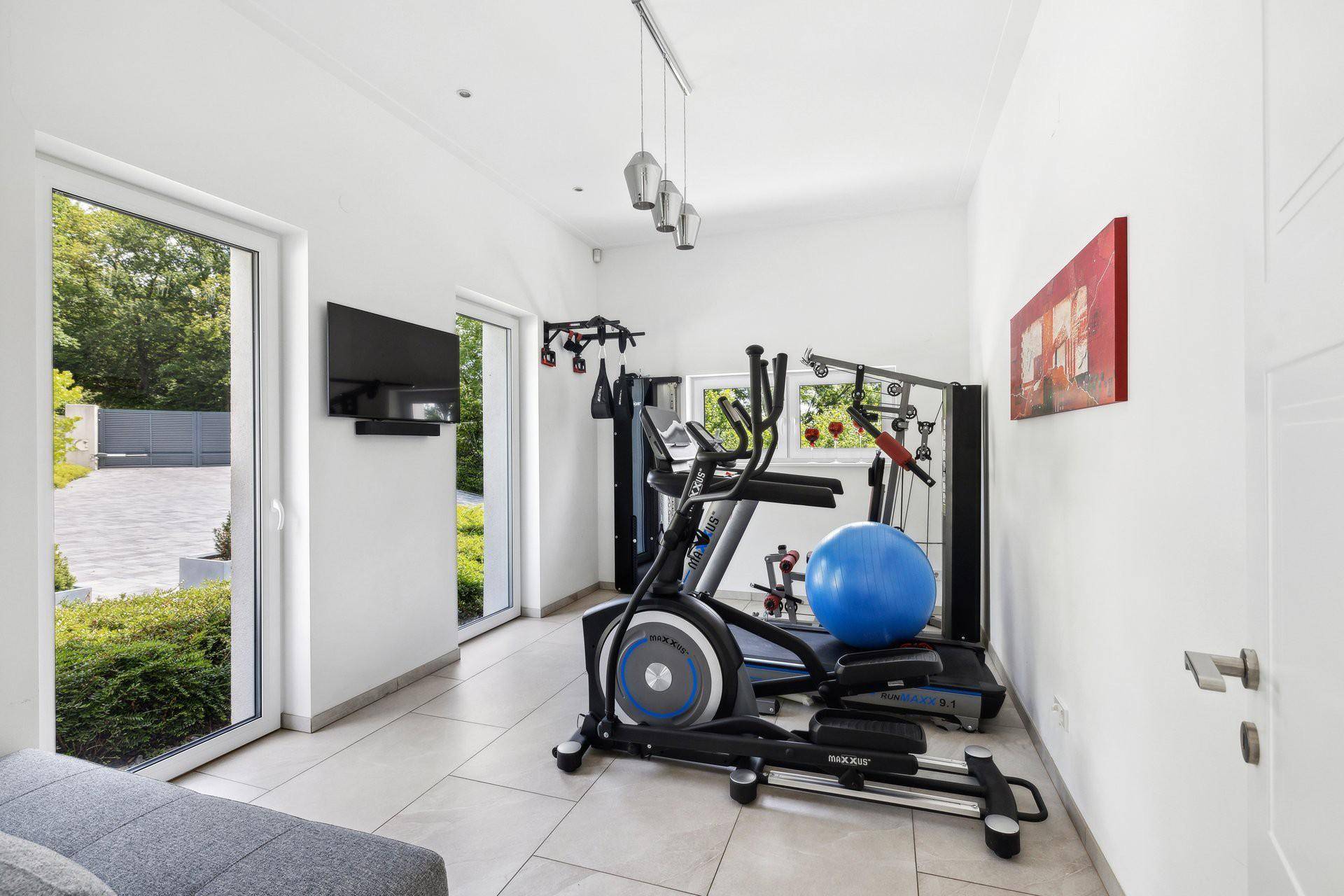 Heller Fitnessraum mit Trainingsgeräten, Fliesenboden und großen Fenstern.