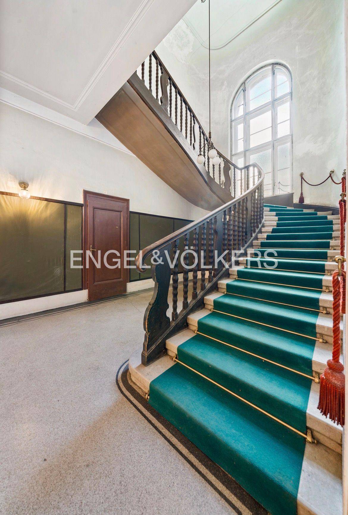 Historisches Treppenhaus mit grünem Teppichläufer, Holzgeländer und großem Bogenfenster.