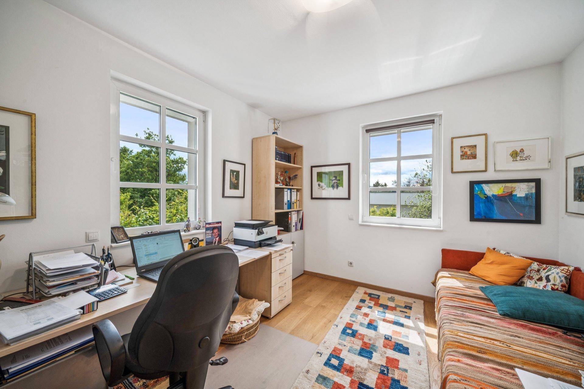 Helles Arbeitszimmer mit Holzfußboden, Schreibtisch und Blick ins Grüne durch große Fenster.