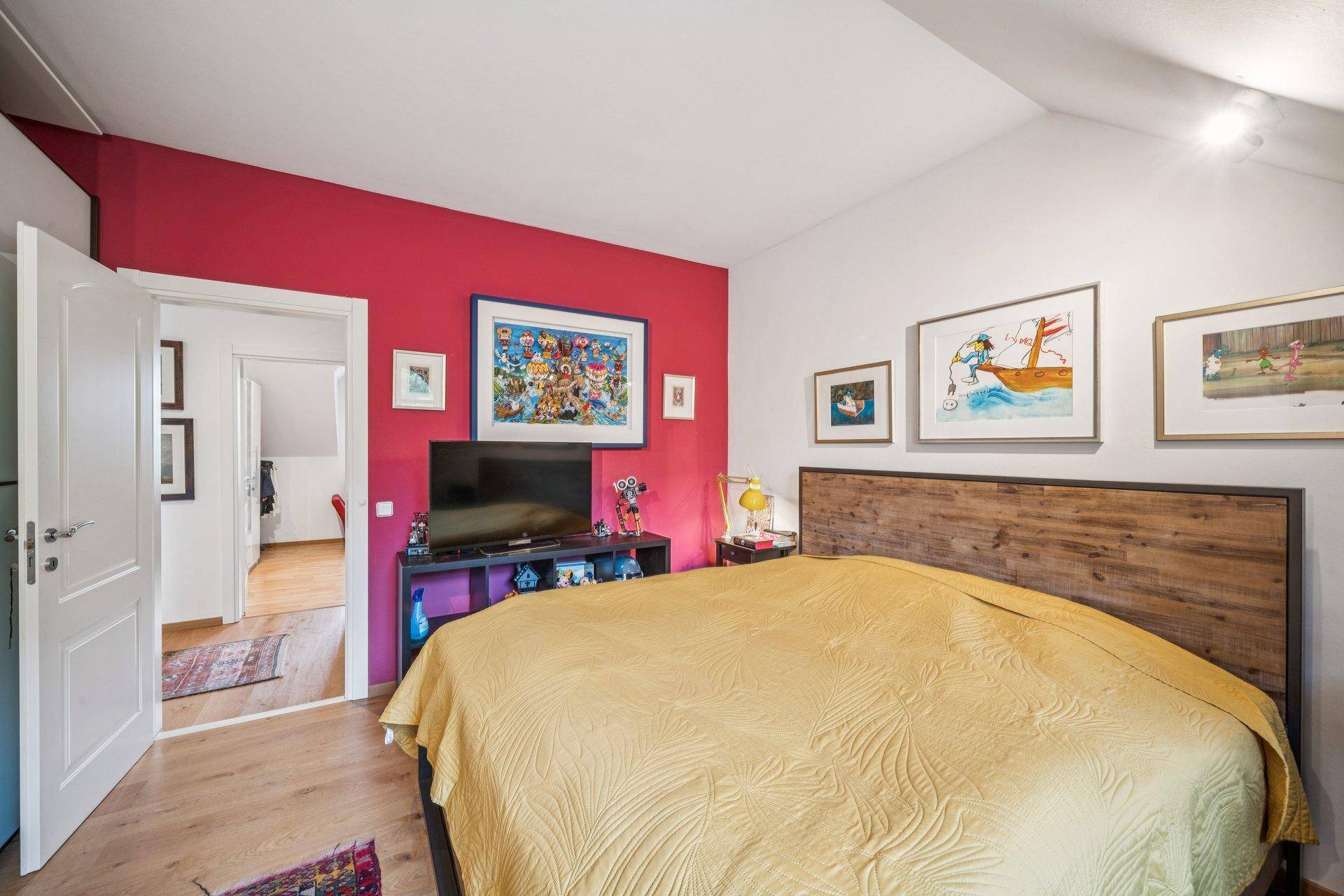 Farbiges Schlafzimmer mit roter Akzentwand, Holzbett und Zugang zu einem weiteren Raum.