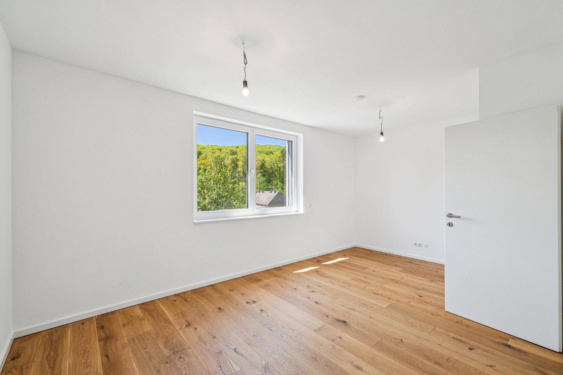 Geräumiges Zimmer mit Holzboden und einem großen Fenster, das einen weiten Blick ins Grüne ermöglicht.
