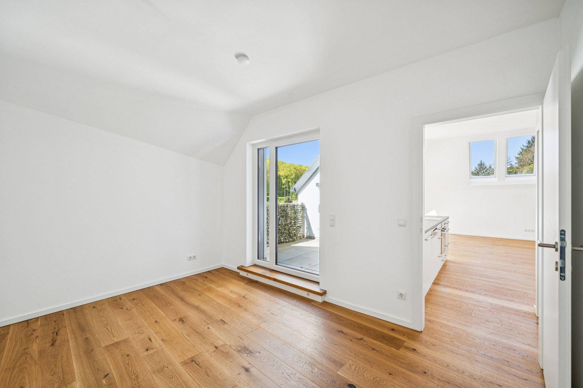 Helles Zimmer mit Holzboden und Zugang zu einem Außenbereich, verbunden mit einem weiteren Raum.