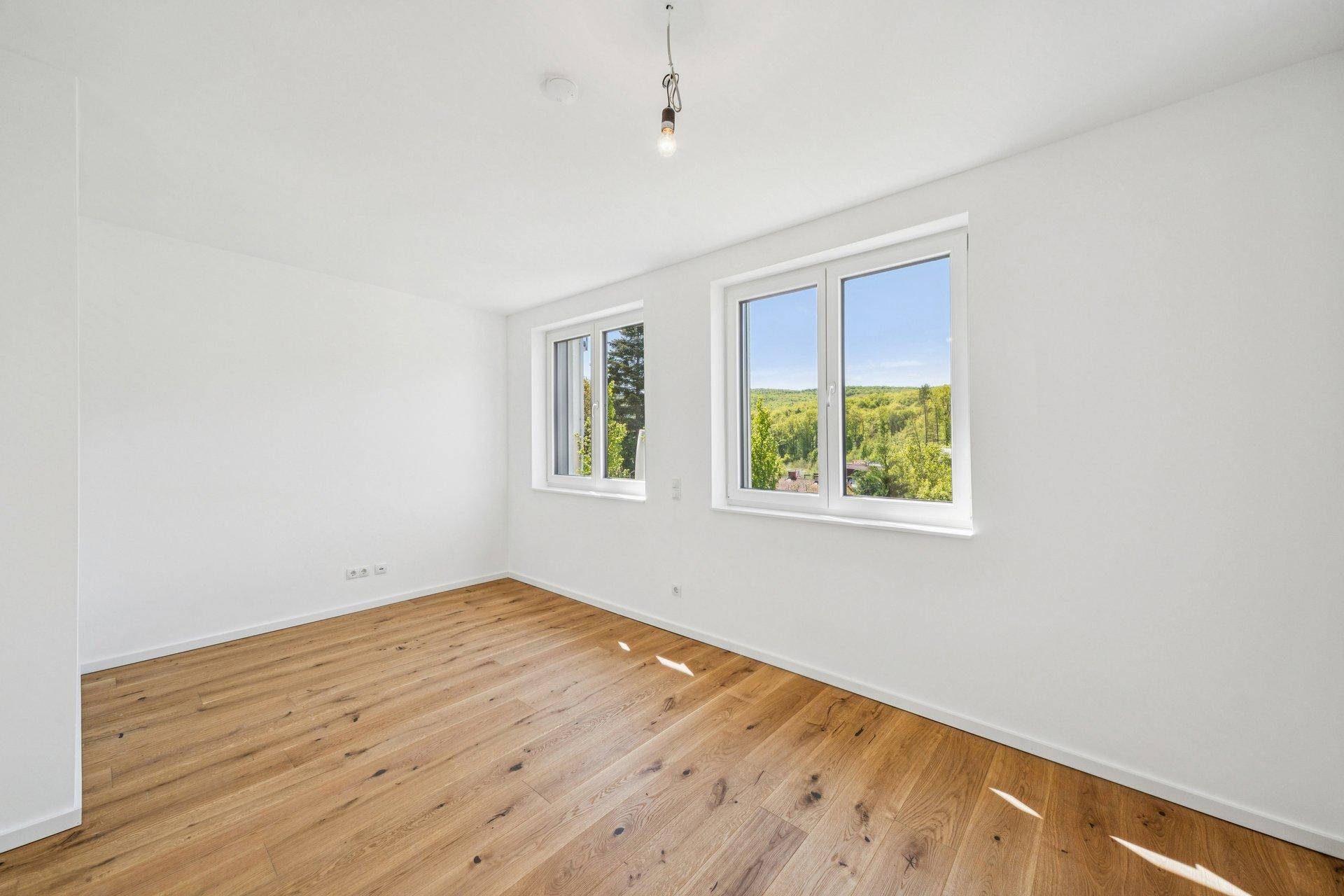 Helles Zimmer mit Holzboden und zwei Fenstern, die einen schönen Blick ins Grüne bieten.